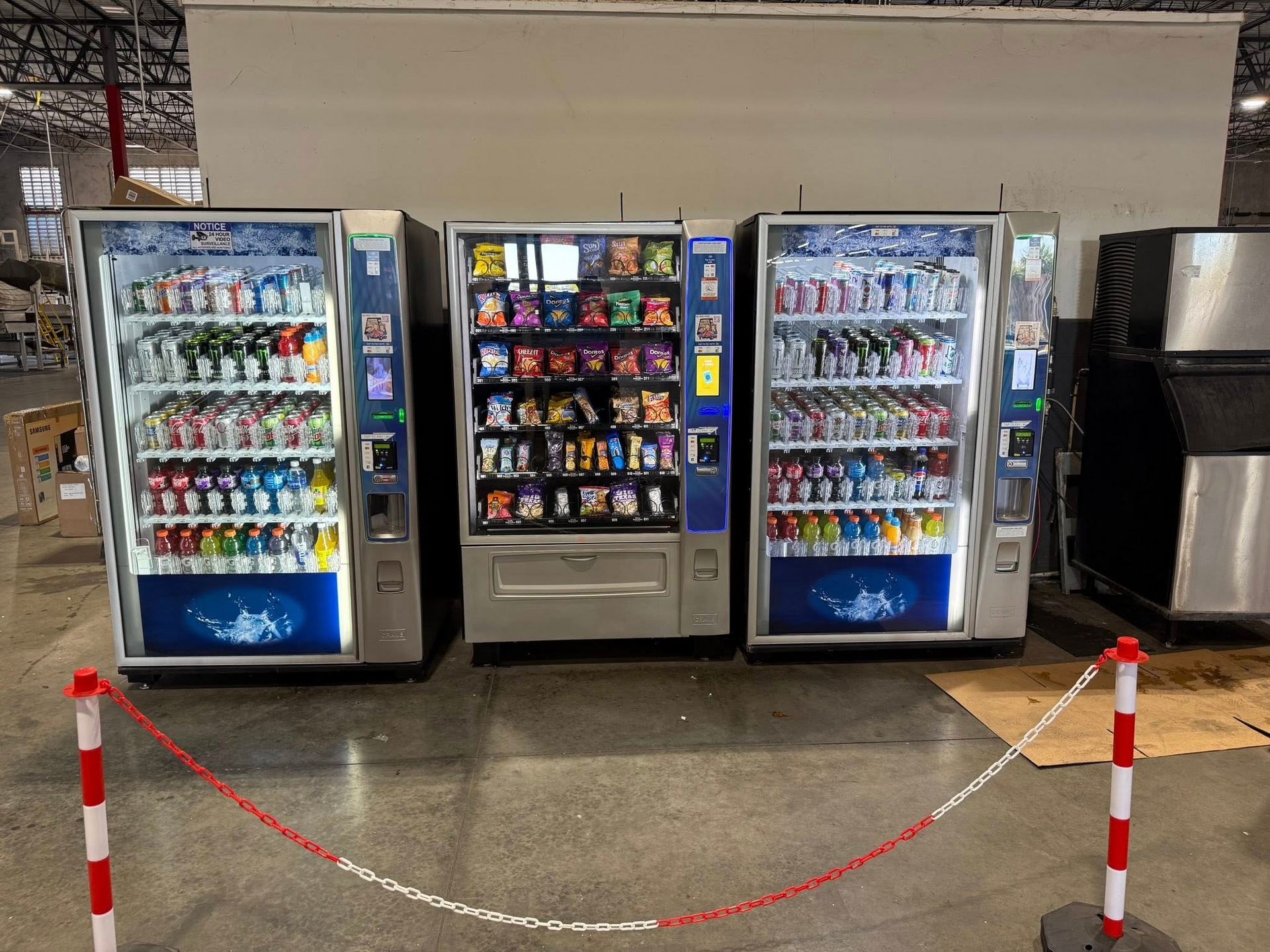 Three vending machines stand side-by-side in a large, open indoor space, fronted by a plastic chain barrier on red poles.