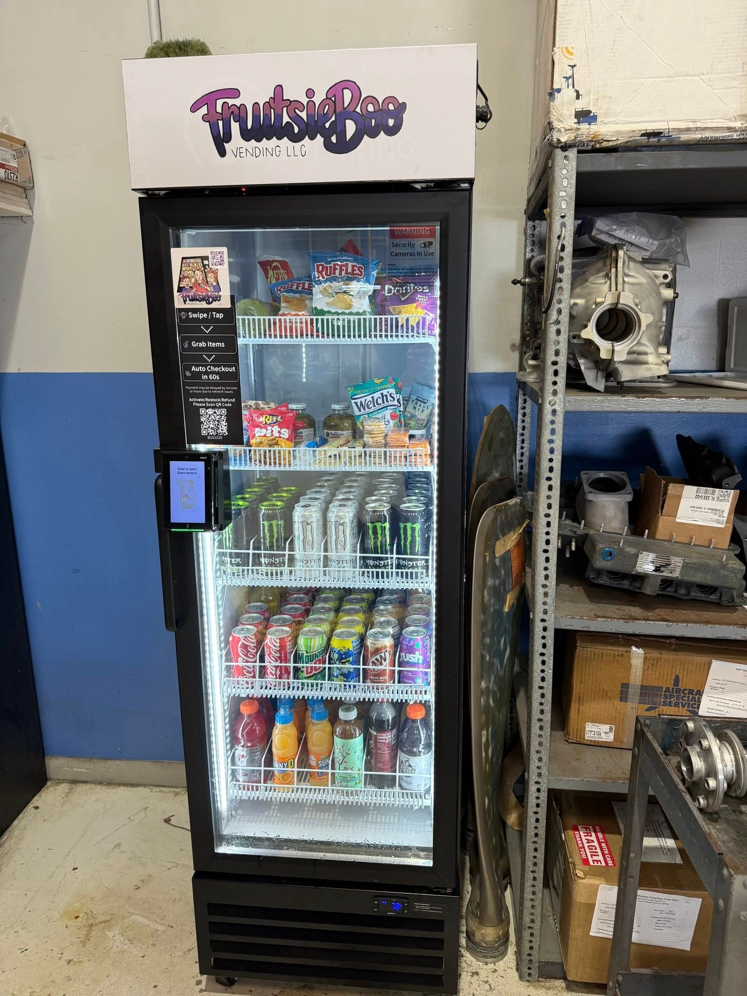 A glass-front FrutsieBoo refrigerator filled with snacks and beverages, located in an industrial workshop setting.