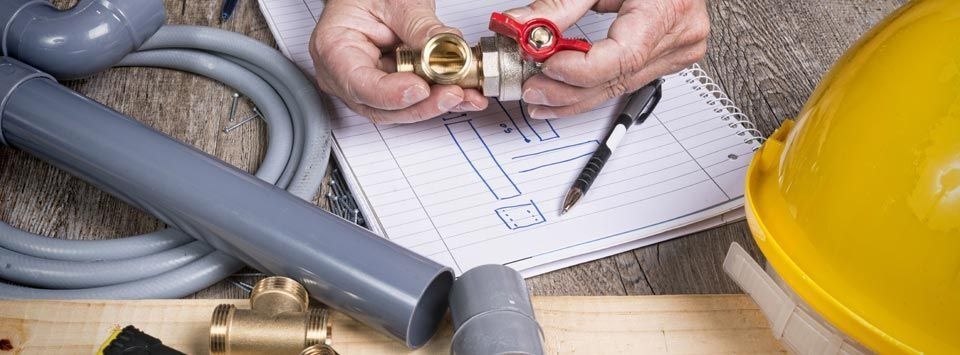 Hands holding plumbing fixture over a blueprint. Various pipes and a yellow hard hat are visible.