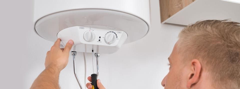 Man repairs a hot water heater, using a screwdriver. The heater is mounted on a white wall.