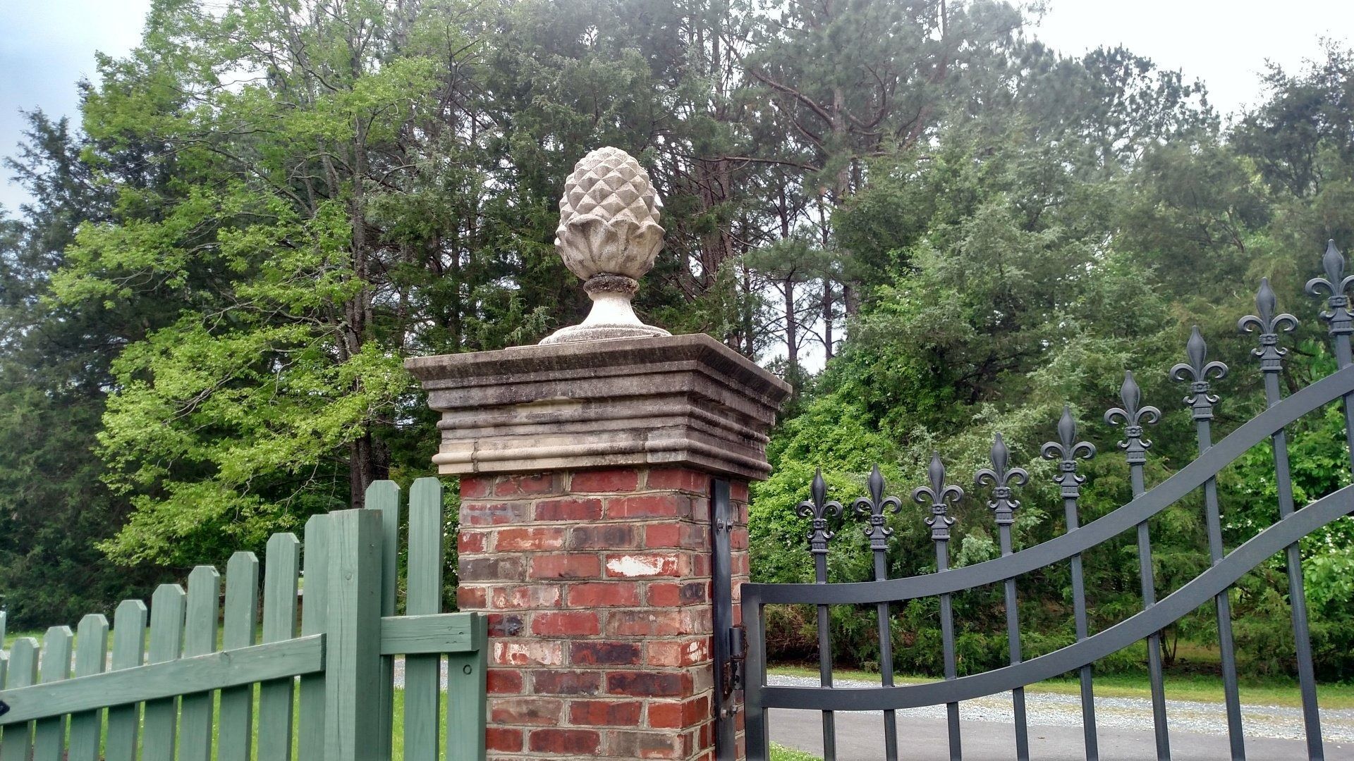 Brick gate pillar with urn finial beside a green metal fence and driveway, with trees in the background