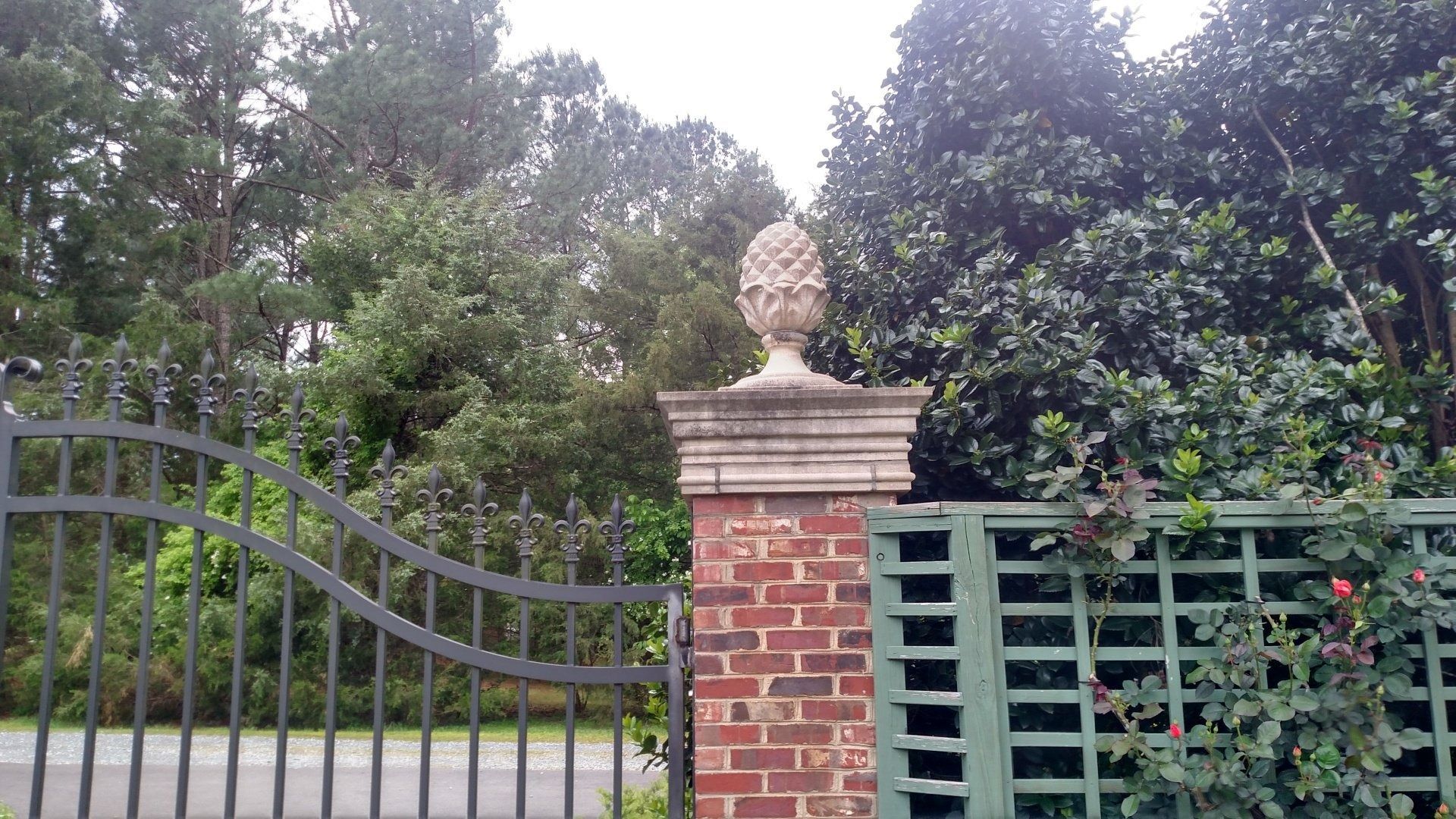 Brick gate pillar with bust beside a metal fence in front of dense green trees