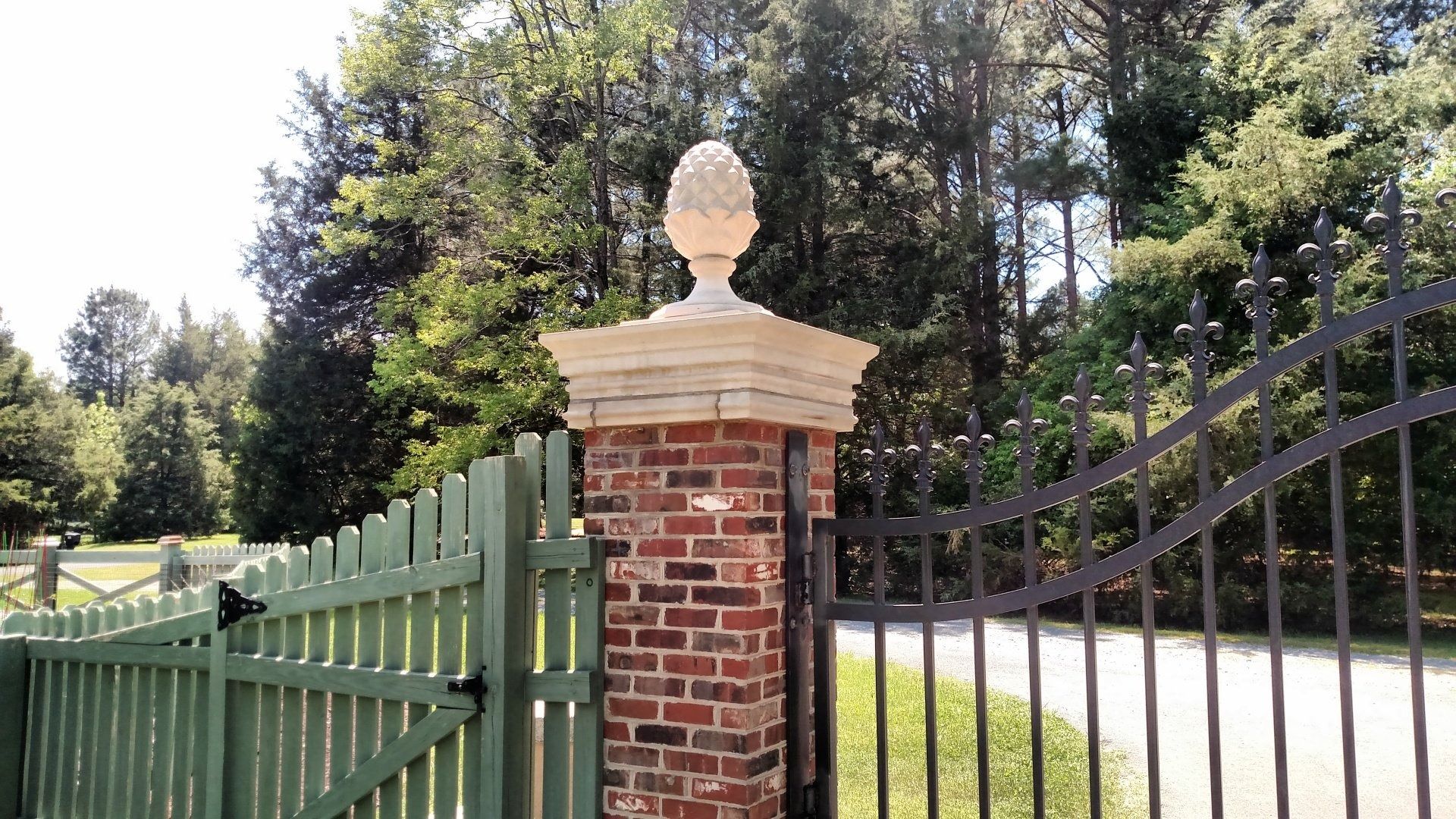 Brick gatepost with white finial beside a green and black wrought-iron driveway gate, with trees behind.