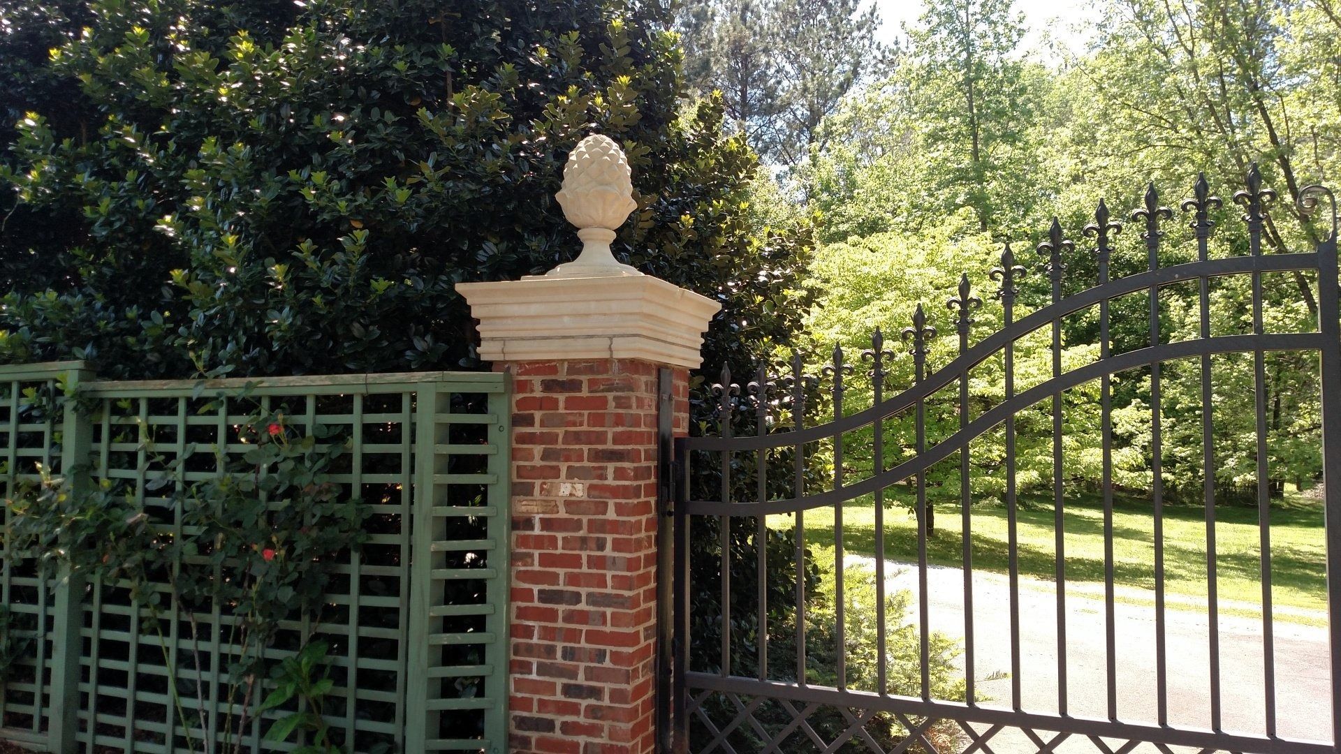Brick gate pillar and black metal gate beside a tree-lined driveway in sunlight