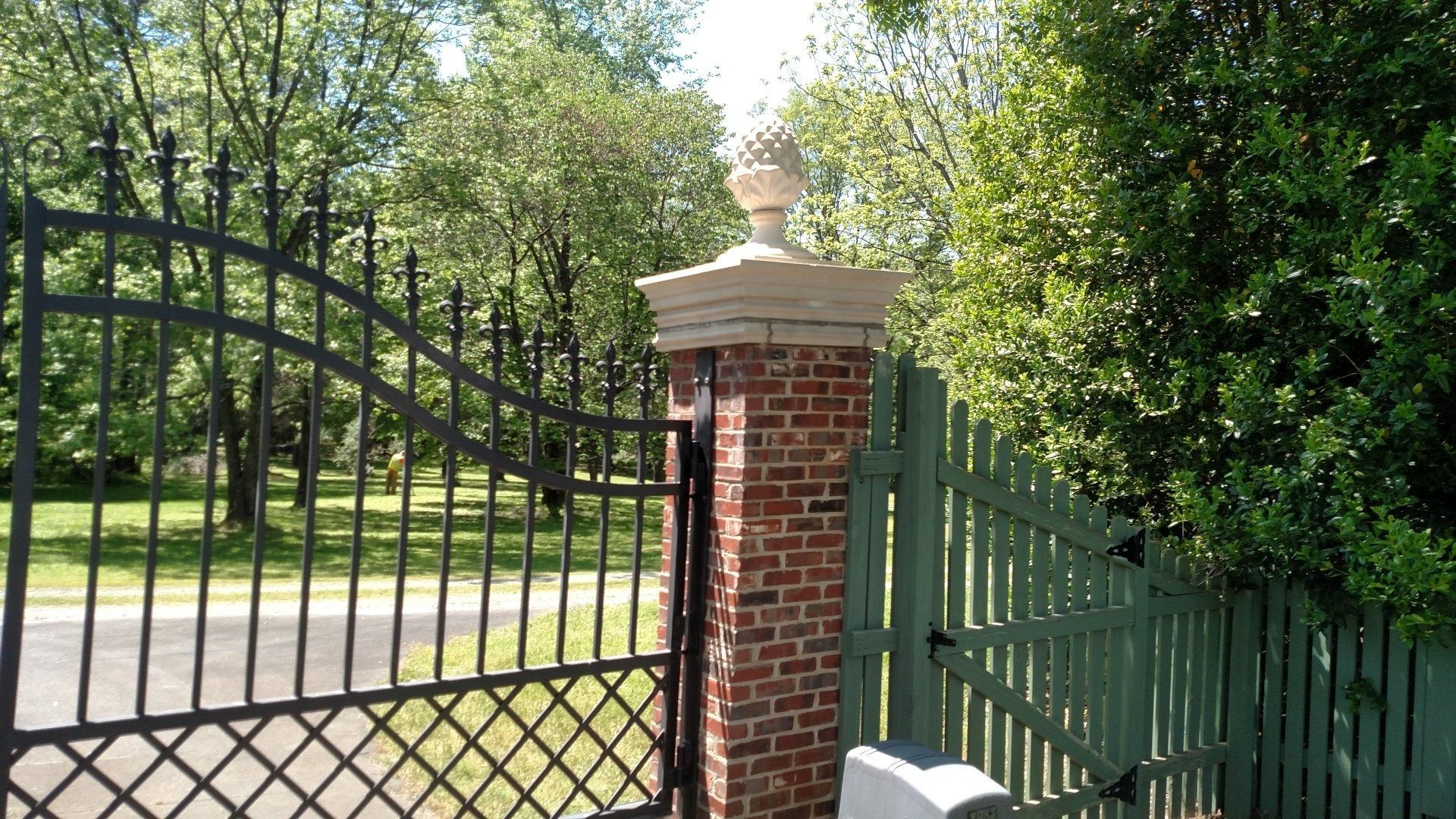 Black wrought-iron gate beside a brick pillar and green hedge by a tree-lined road