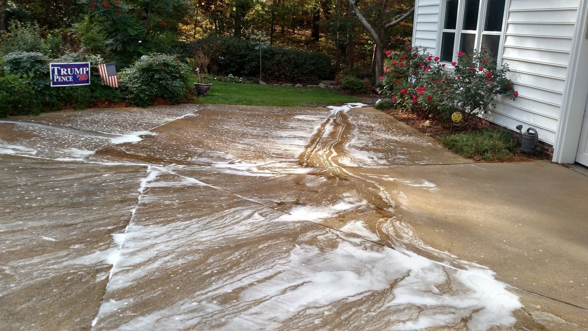 Foamy runoff water flowing across a driveway beside a house and garden in the rain