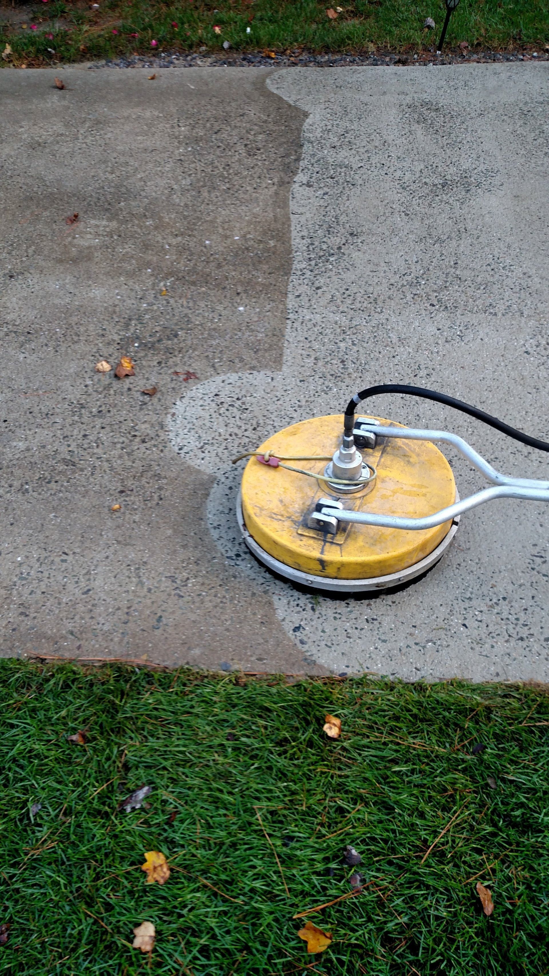 Yellow pressure washer on a concrete walkway, spraying water and cleaning a gray patch near the grass edge.