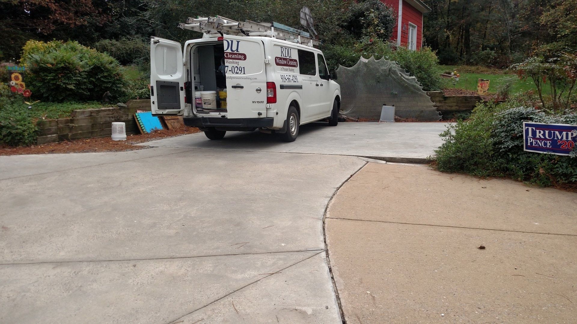 White utility van parked in a driveway beside a brick house and shrubs, with campaign signs nearby