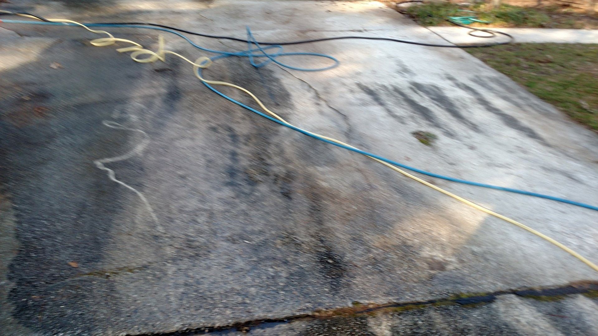 Driveway pavement with dark stains and a blue hose stretched across it in bright sunlight