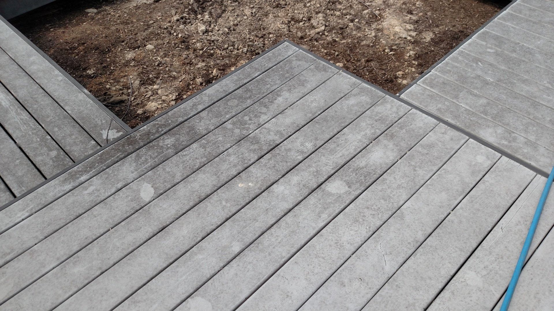 Gray wooden deck boards arranged in a geometric pattern beside a dirt patch and blue cable