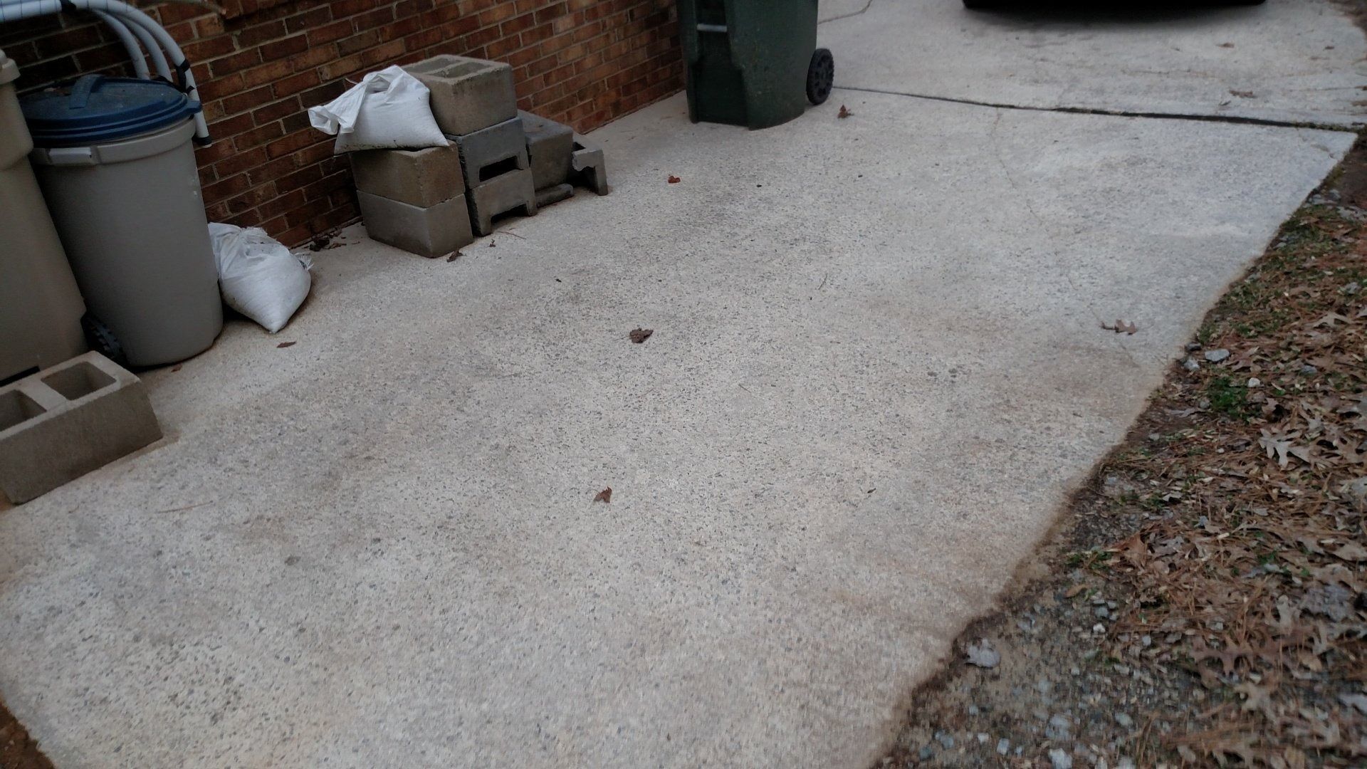 Outdoor concrete driveway with a gravel edge, trash bins, and stacked boxes near a house