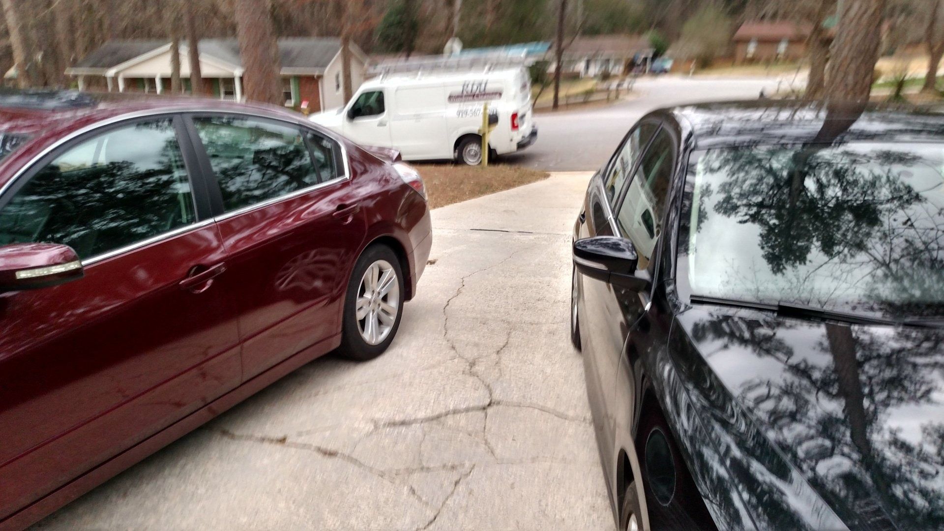 Two parked cars framing a cracked driveway with a white van and trees in the background