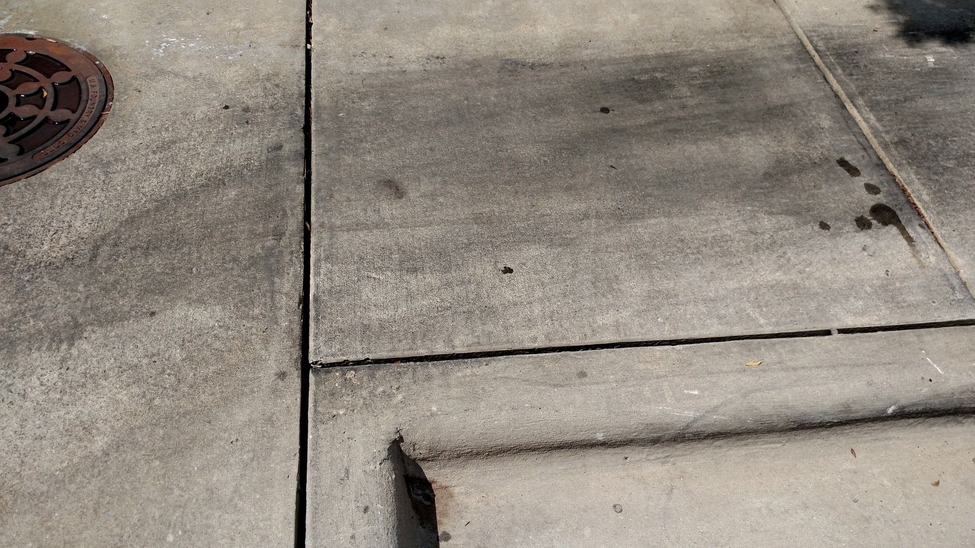 Cracked concrete sidewalk with a curb, drain grate, and small dark stains