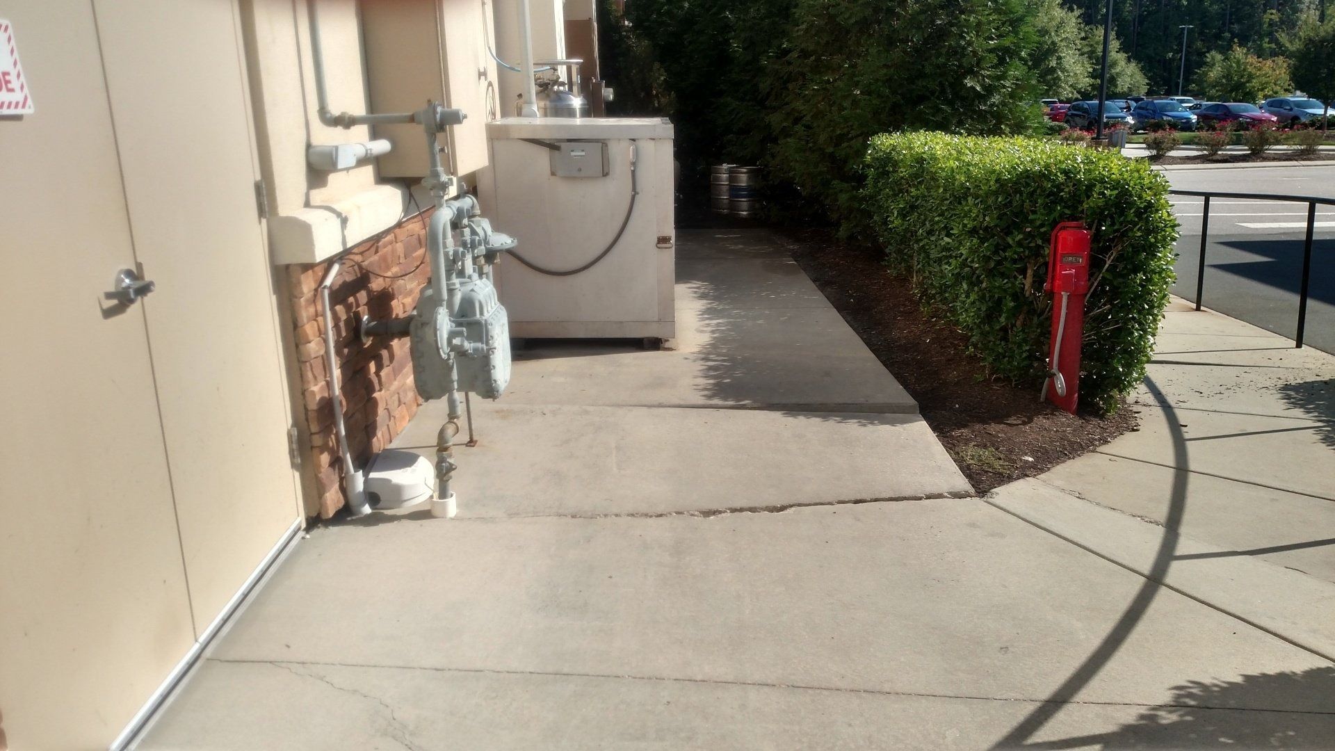 Sidewalk beside a building with a trash bin, shrubs, and a red bollard near a parking area.