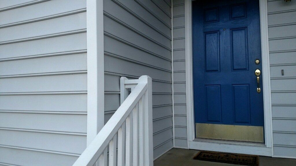 Blue front door with brass knob beside white siding and porch railing.