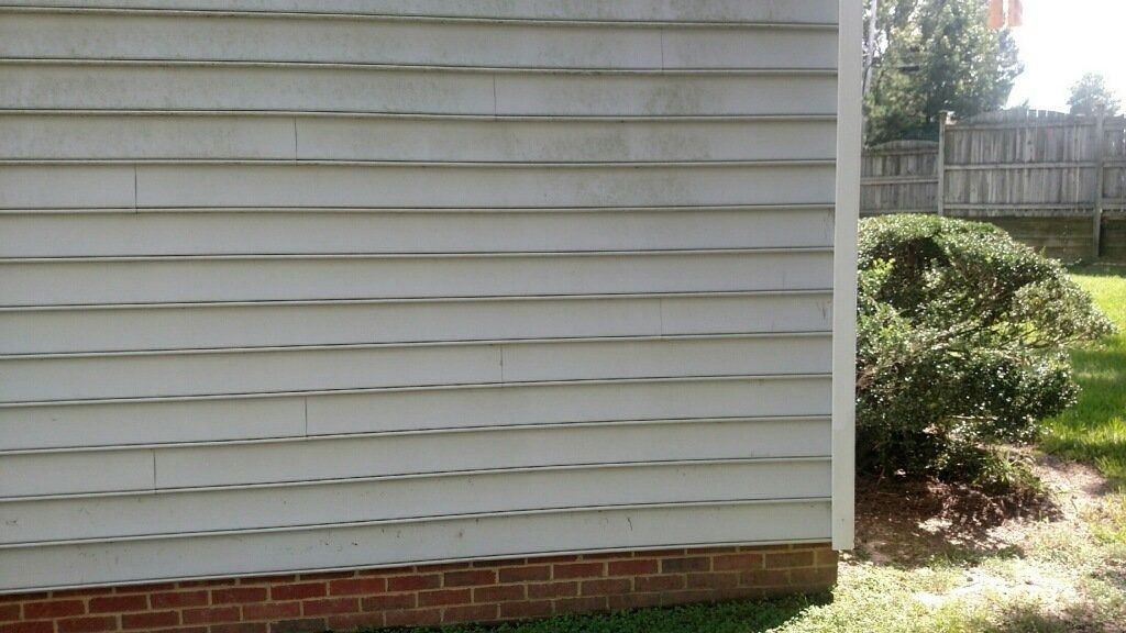 Side of a house with white siding, brick foundation, and a shrub beside a grassy yard.