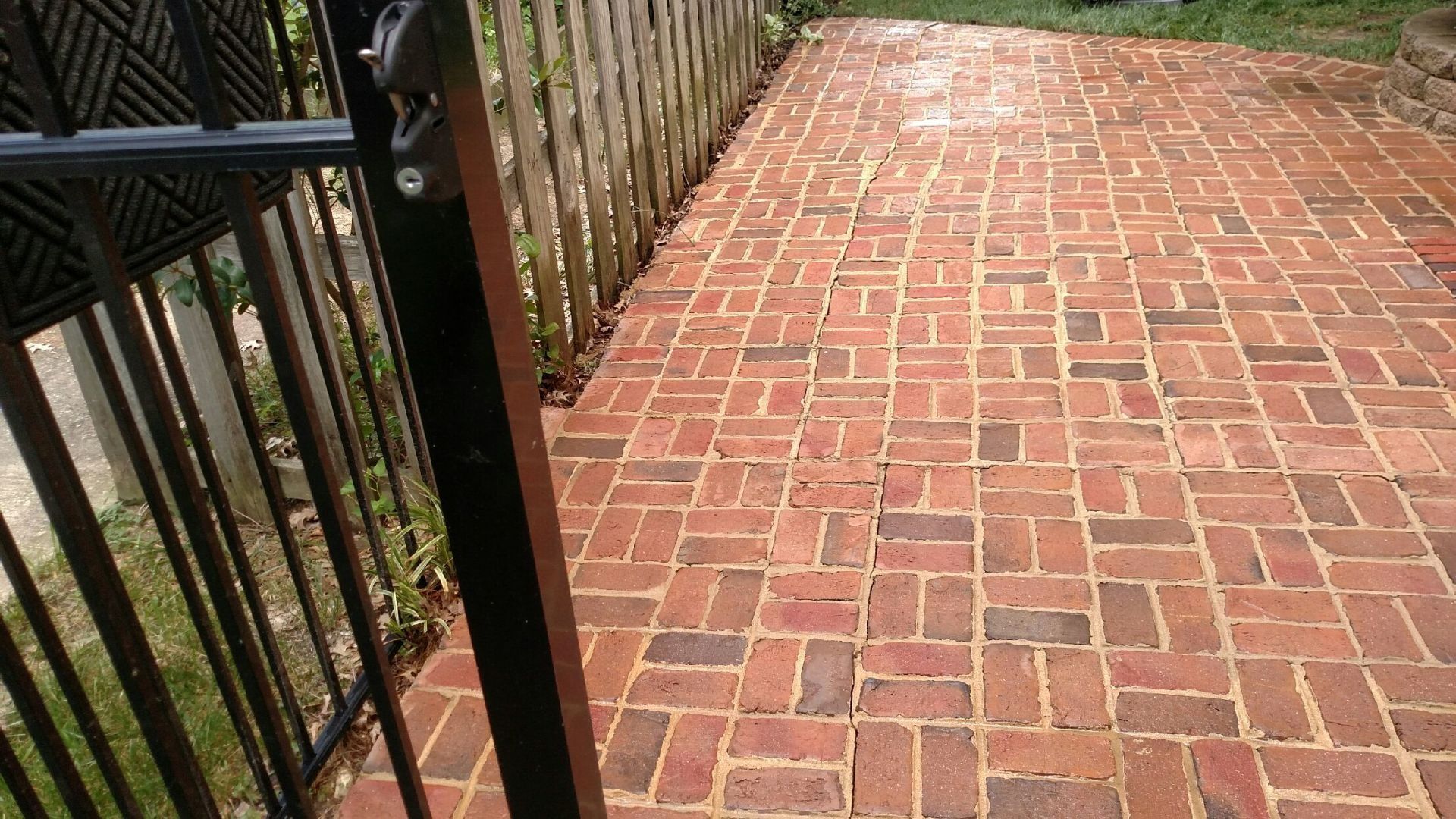 Brick-paved walkway beside a black metal railing and wooden fence, with greenery along the left side.