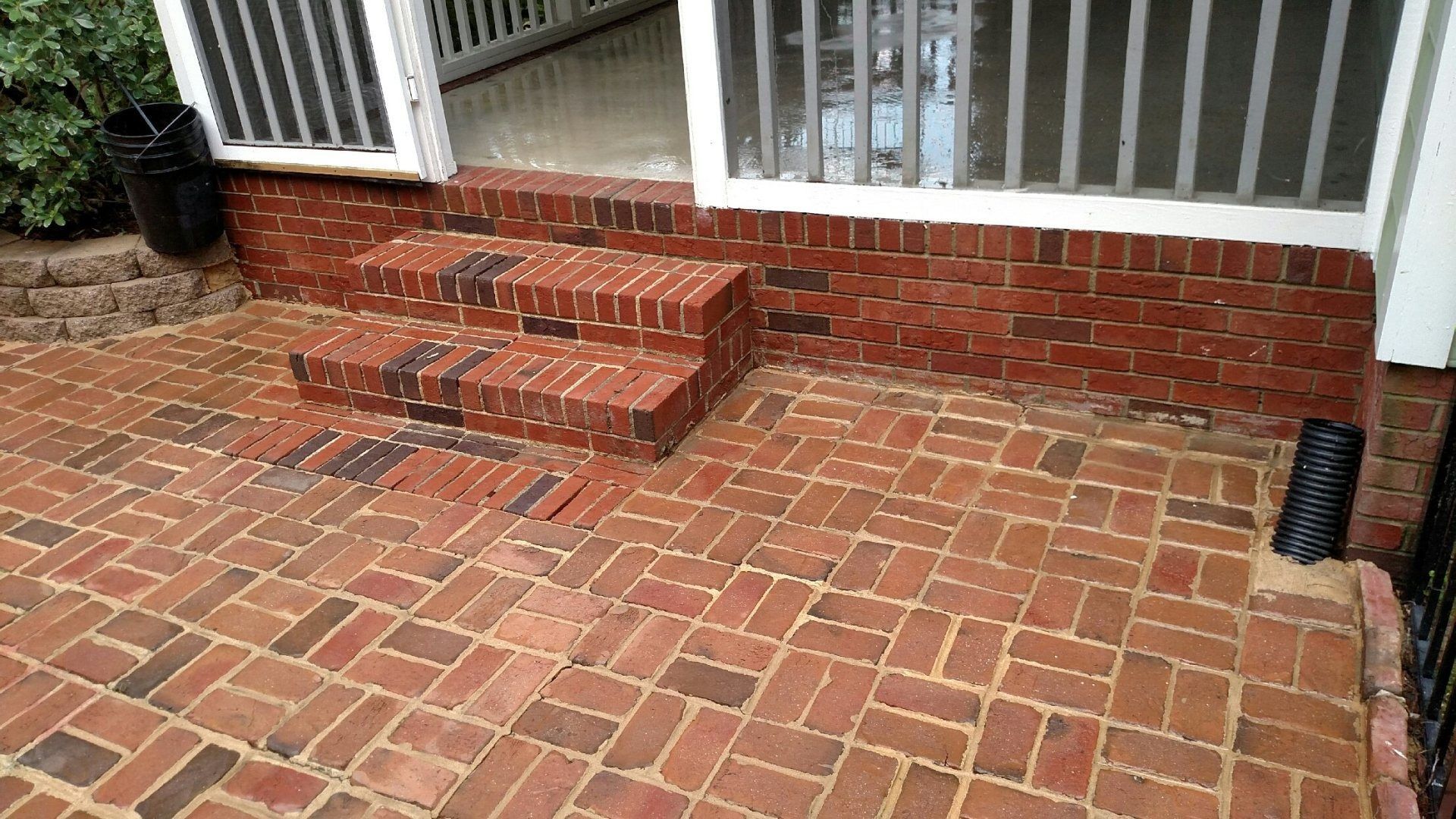 Brick front porch with three steps, white railing, and patterned brick patio, wet from rain.