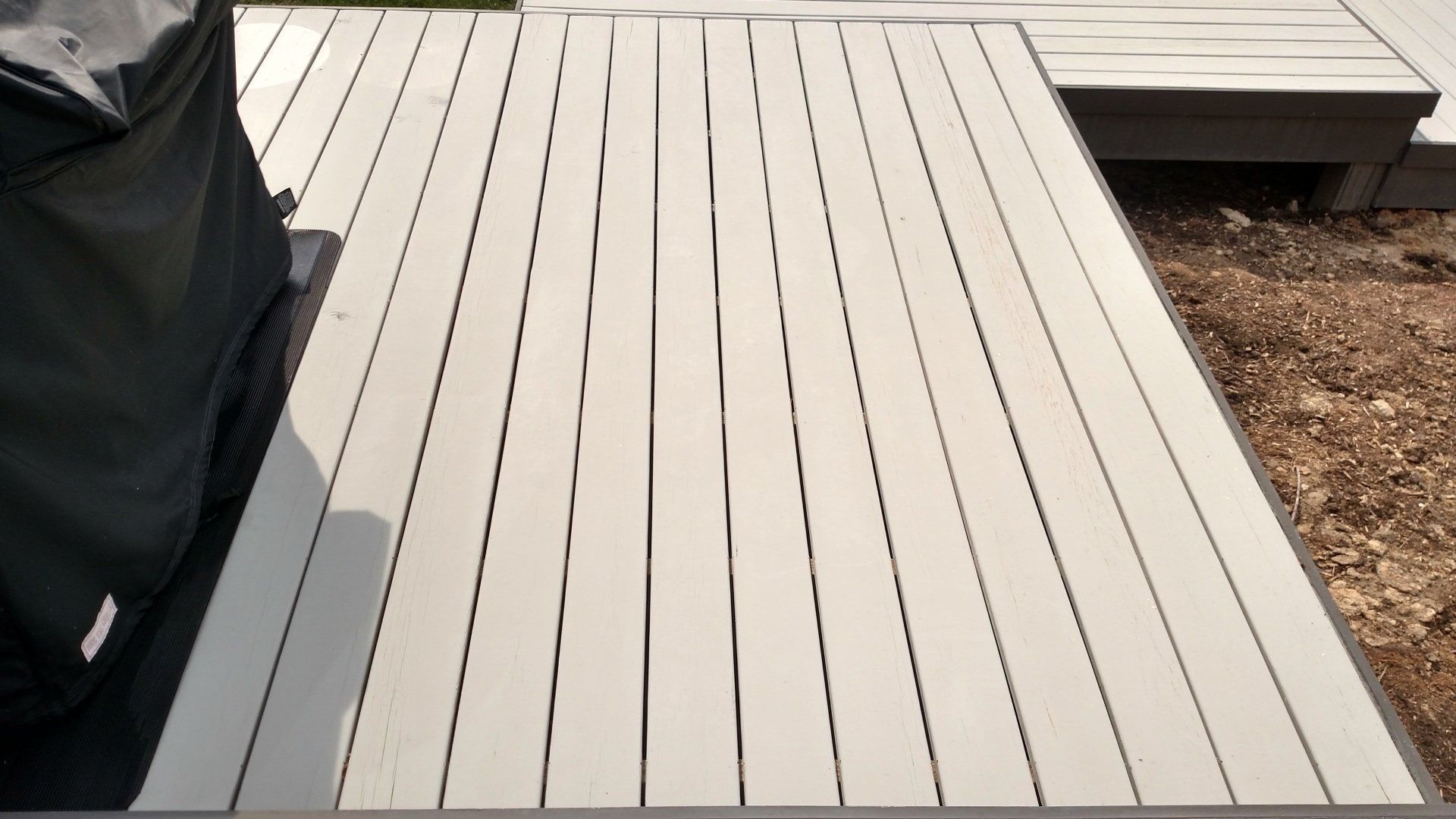 Light gray wooden deck with parallel planks beside a covered grill and a small step platform.