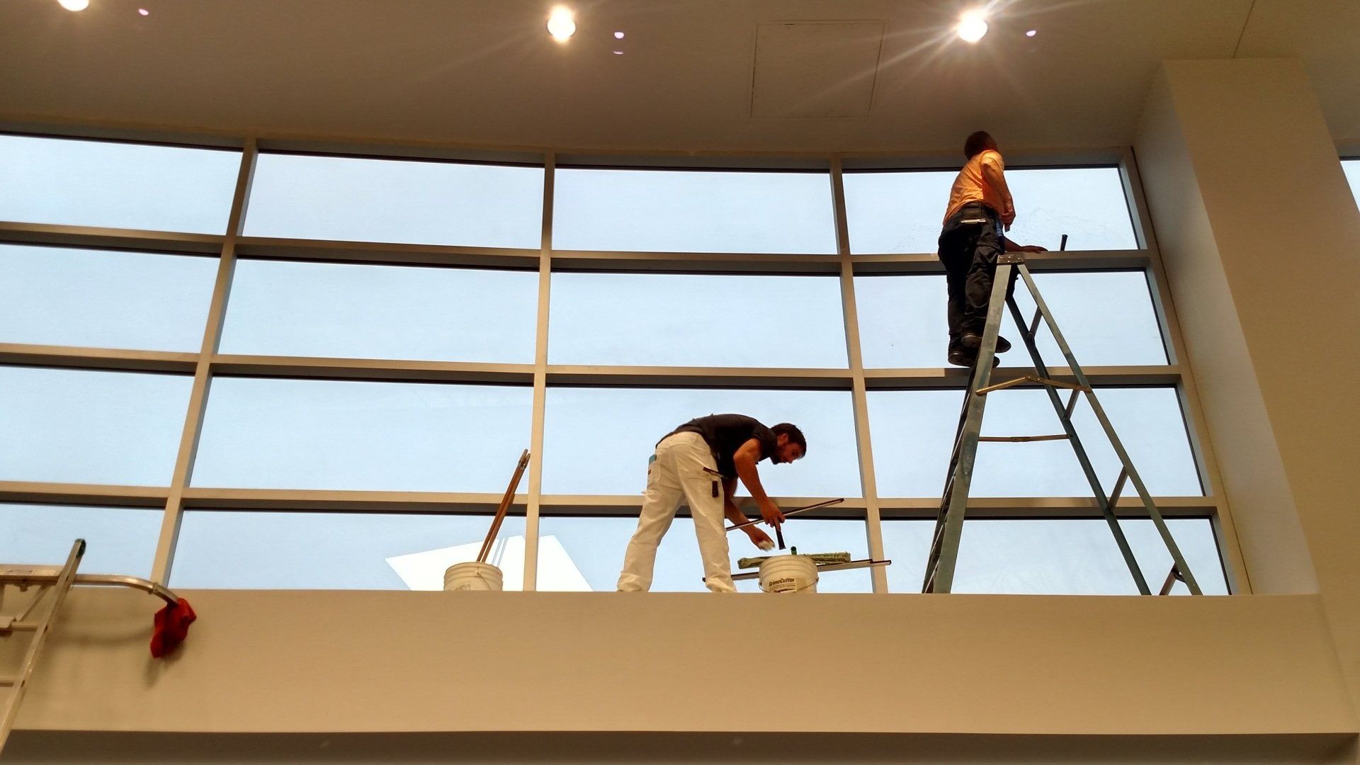 Two workers cleaning large windows on a high interior ledge, one on a ladder