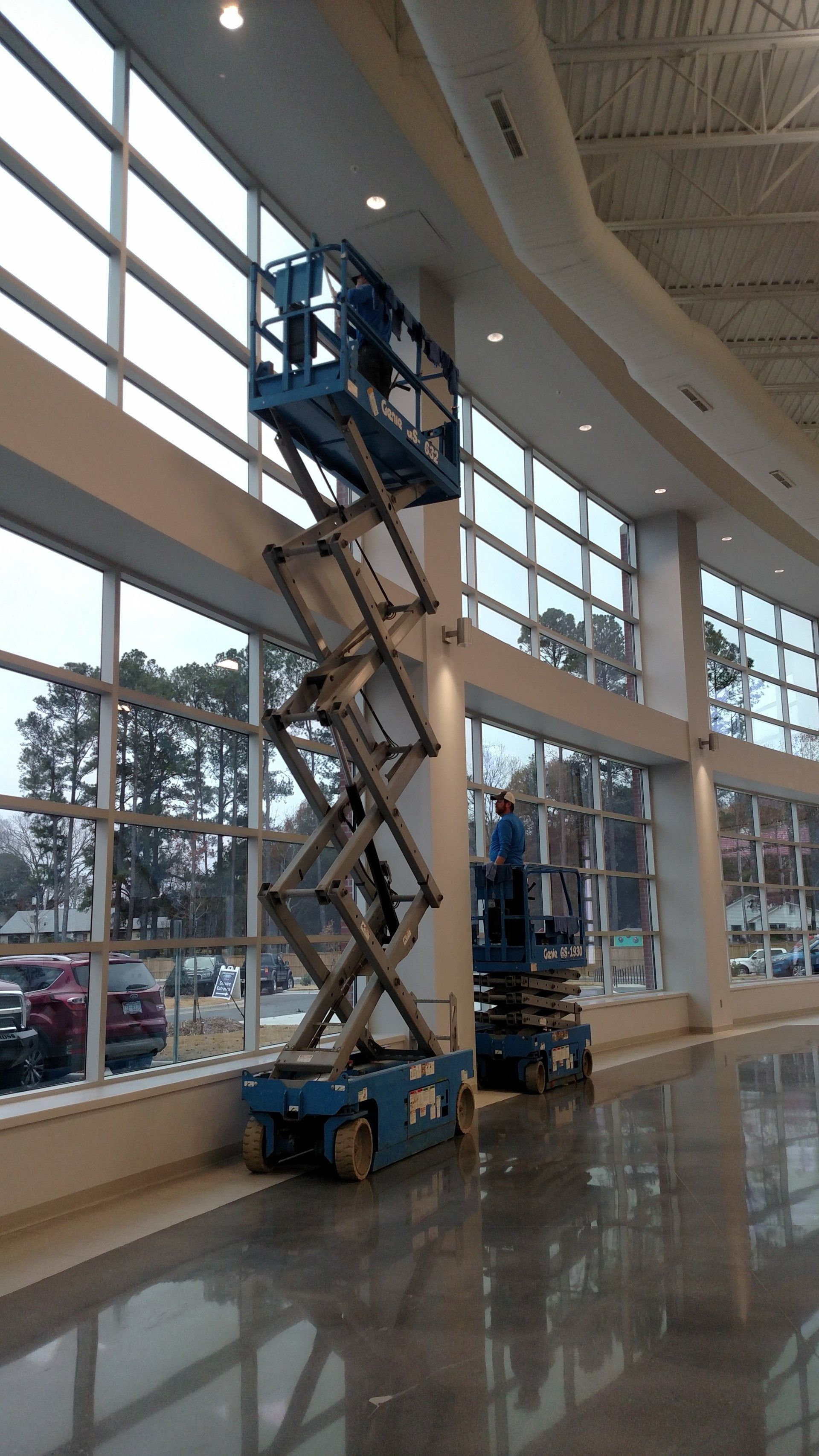 A scissor lift with workers raised in a bright lobby with large windows and a polished floor.