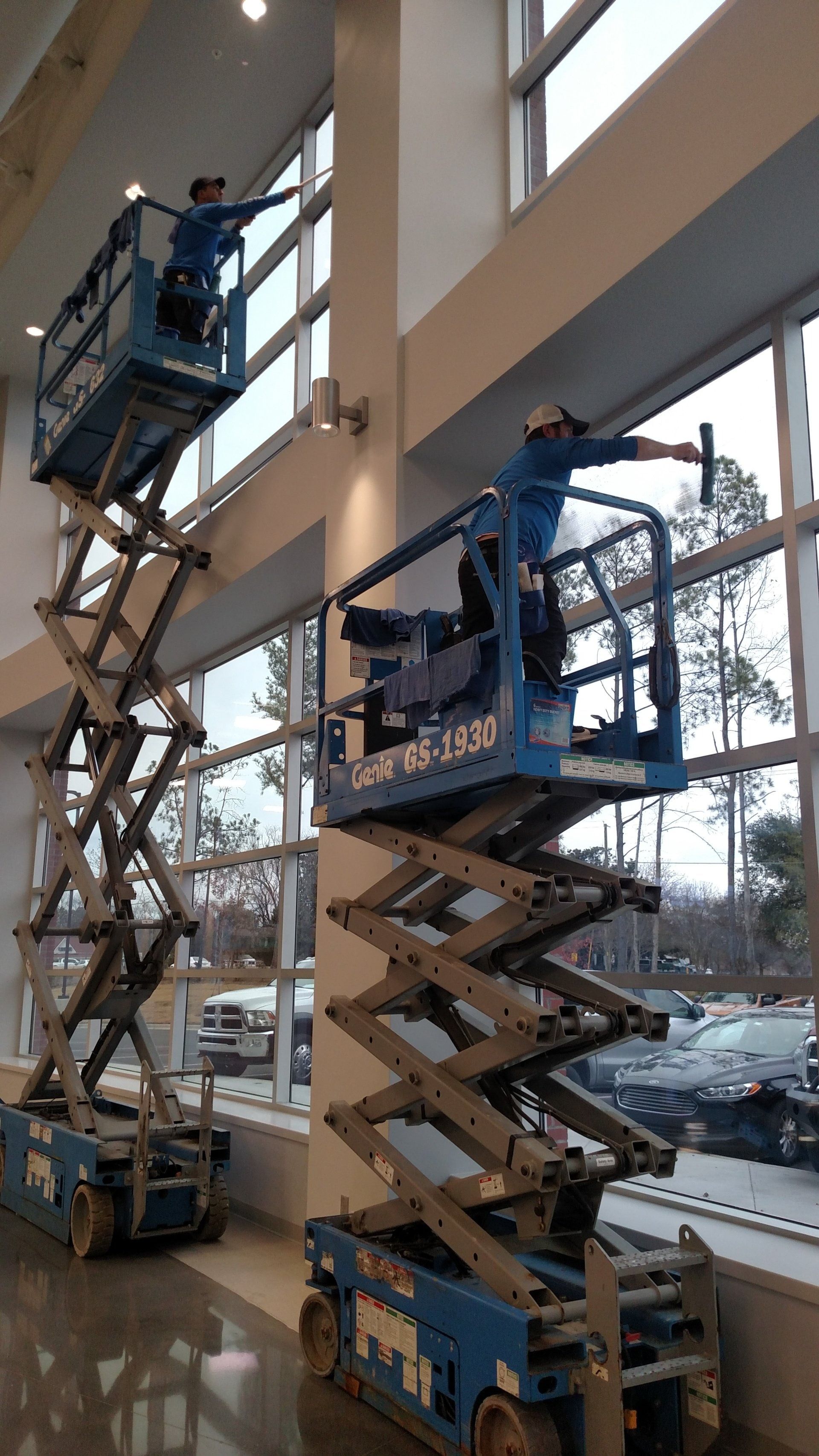 Two blue scissor lifts extend workers near tall windows inside a bright building.