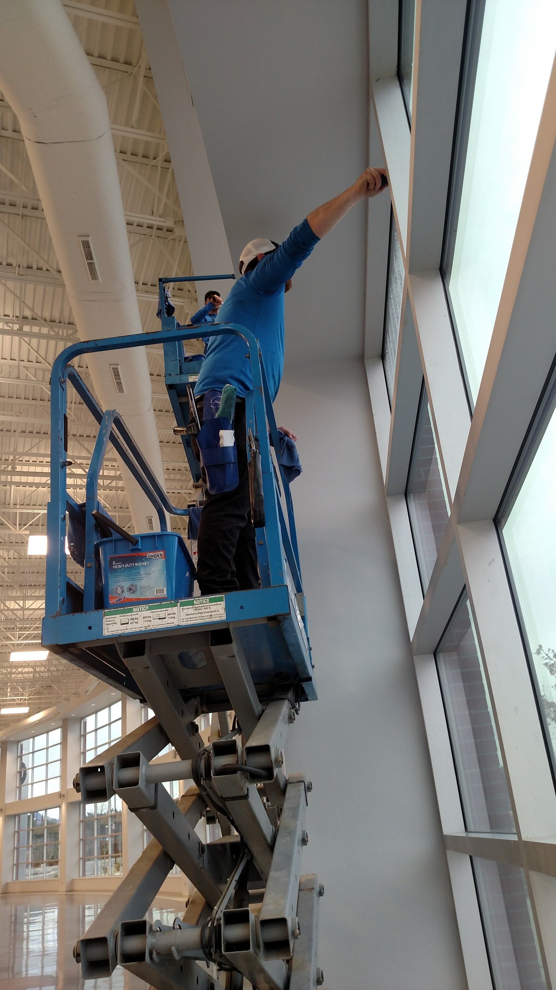 Worker in blue uniform on a lift cleaning tall windows inside a bright building.