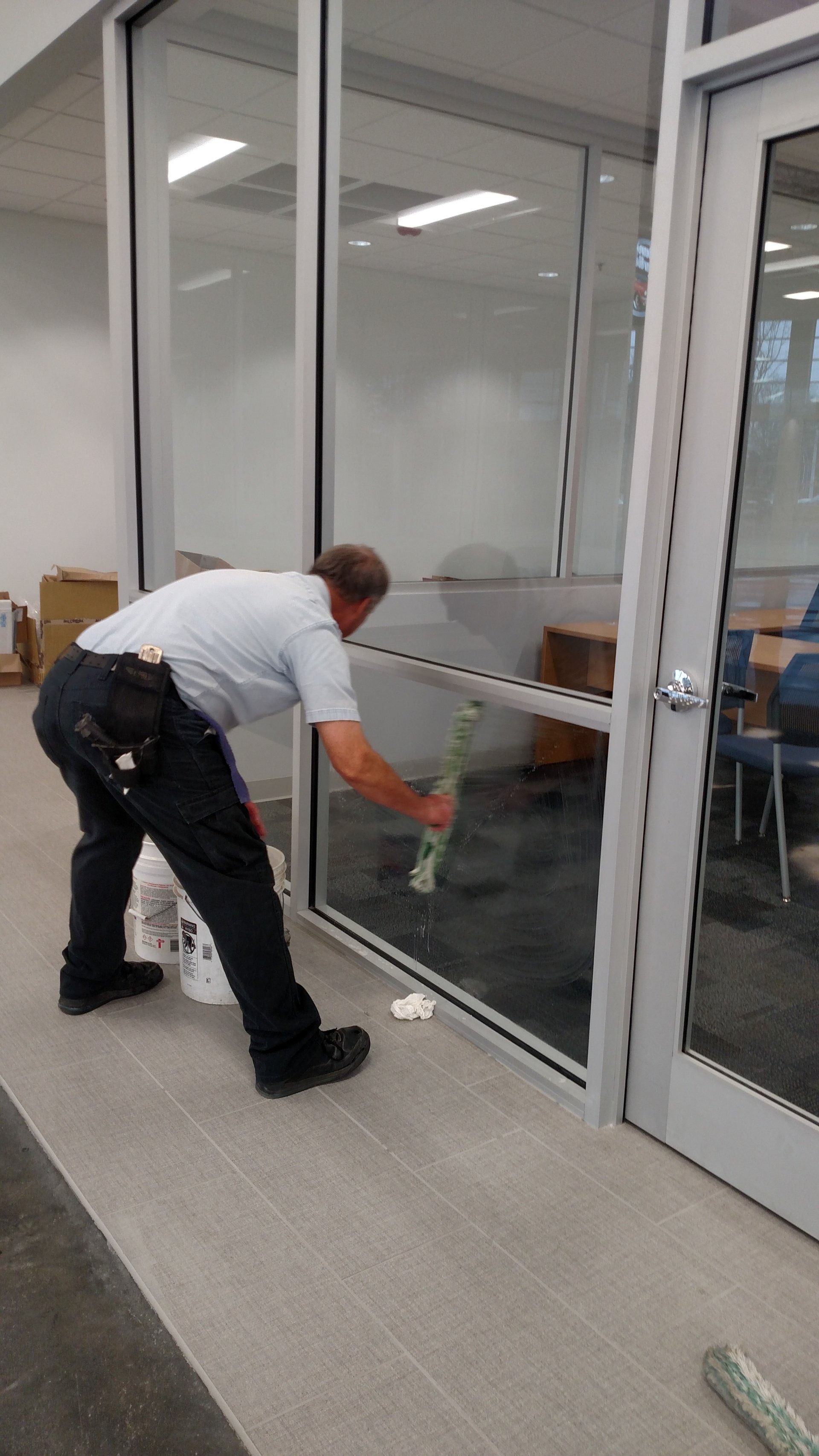 Man cleaning a glass office partition with a cloth in a bright indoor hallway.