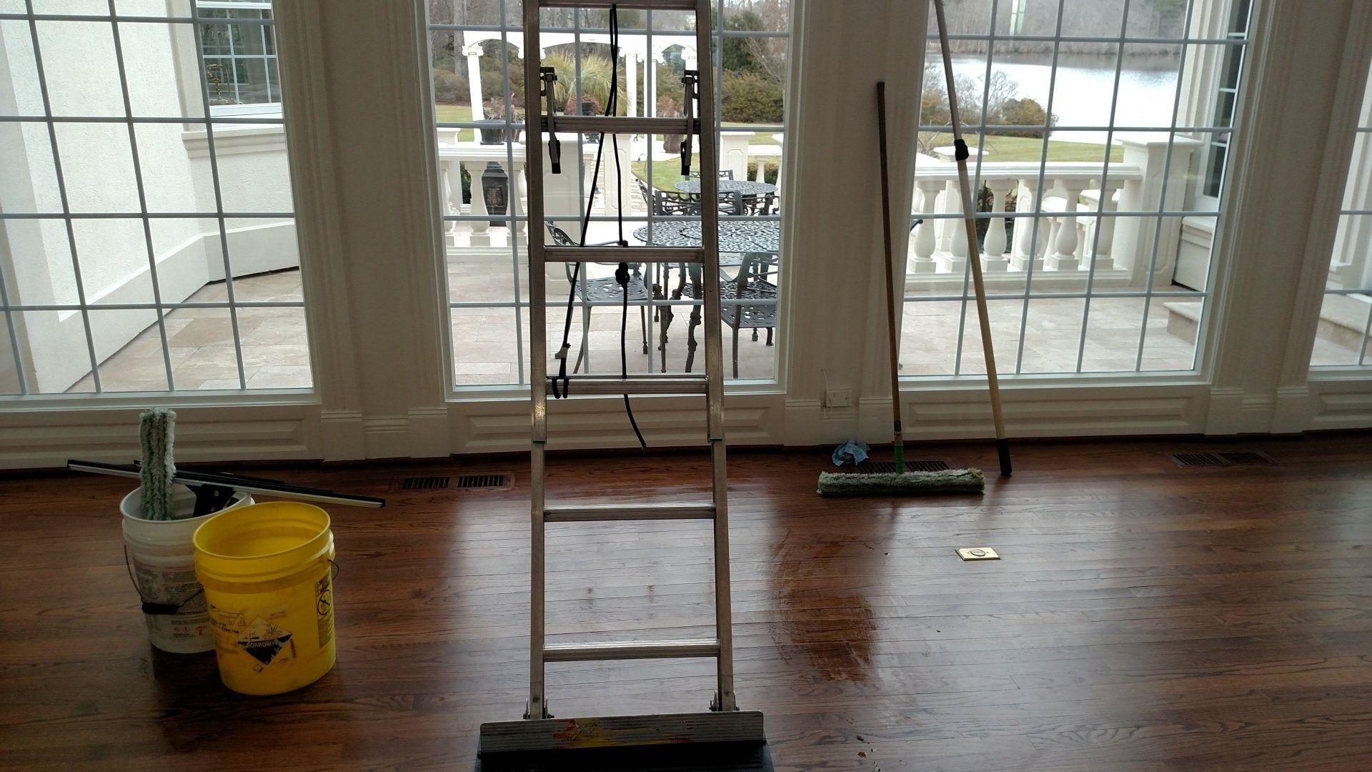 Ladder on a hardwood floor beside a yellow bucket, in front of large windows during cleaning or painting work