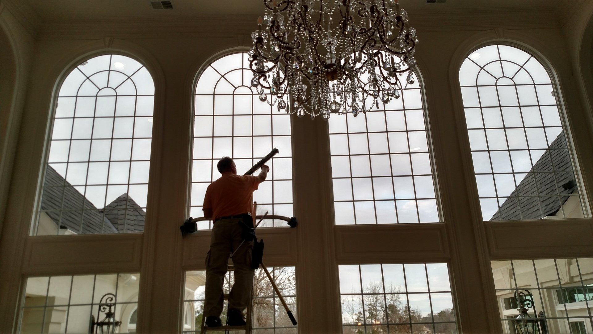 Worker cleaning tall windows in a large hall beneath a chandelier
