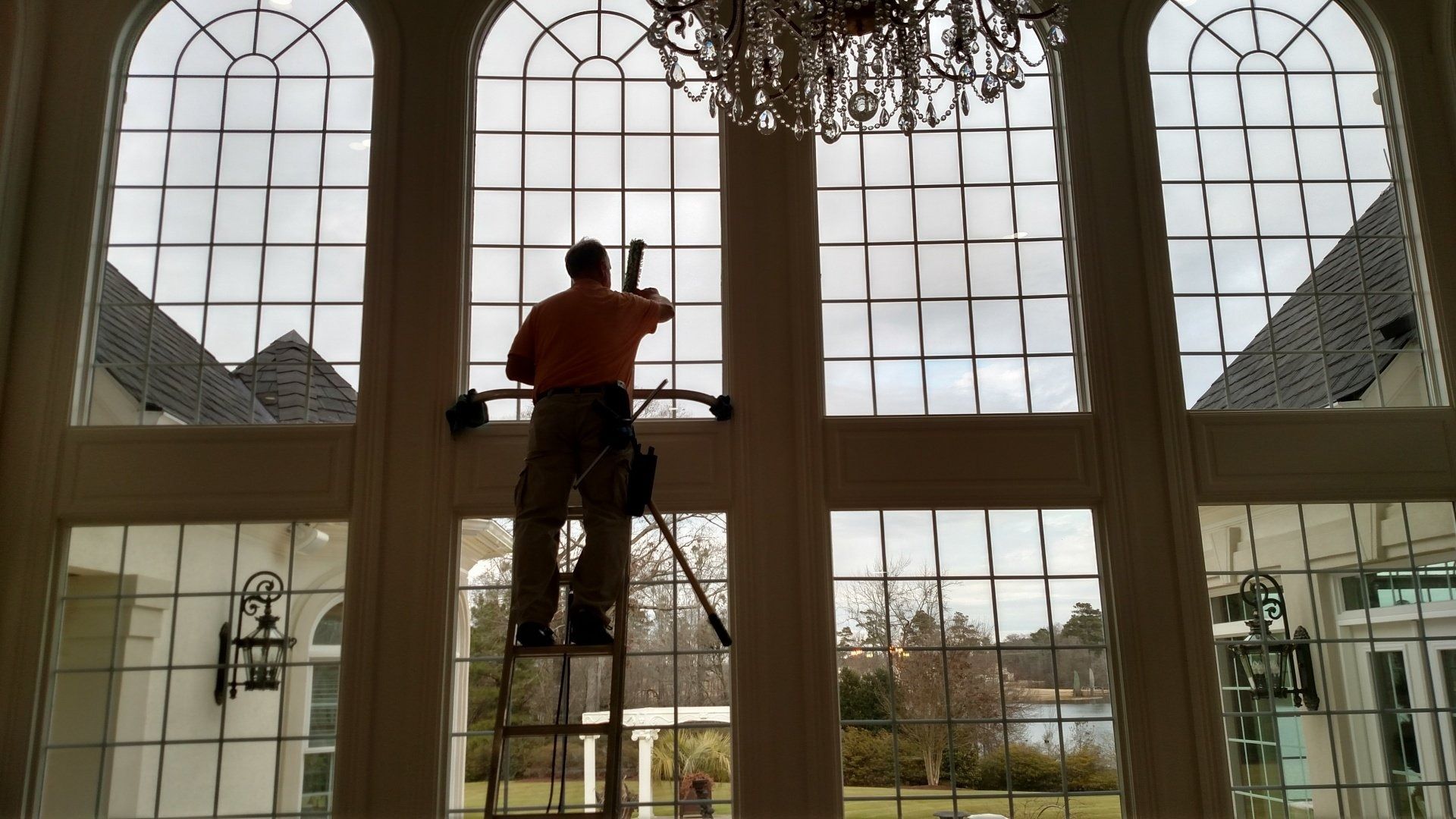 Person on a ladder cleaning large arched windows in a bright room, backlit by daylight