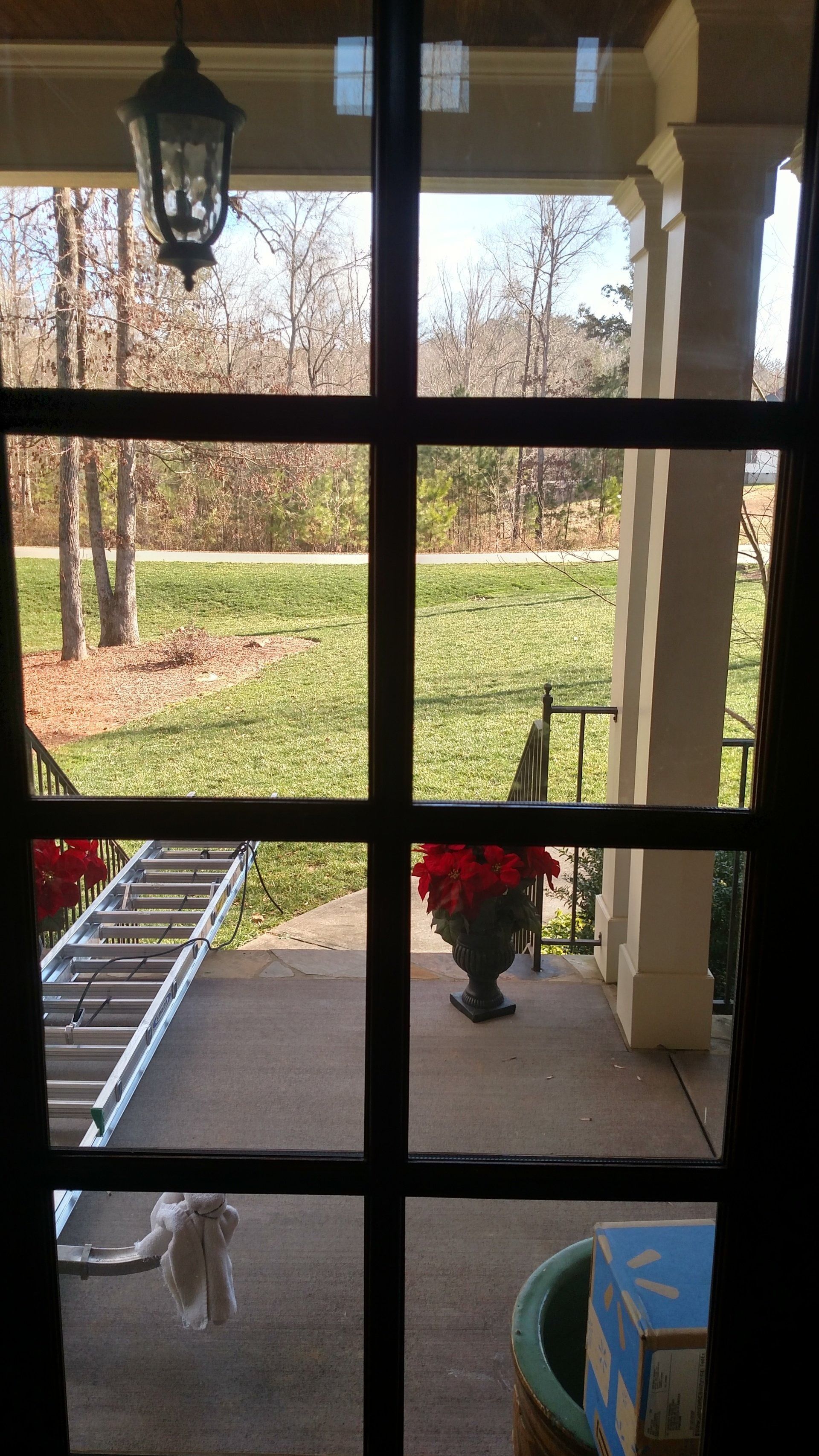 Front porch view through a window grid, with a ladder, hanging lantern, potted plant, and yard beyond.