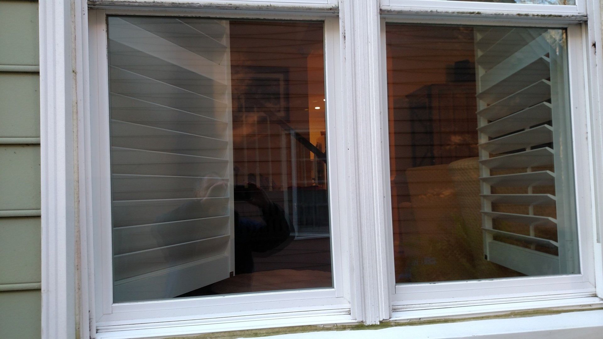 Closed white window with slatted shutters, reflecting a room inside at dusk.