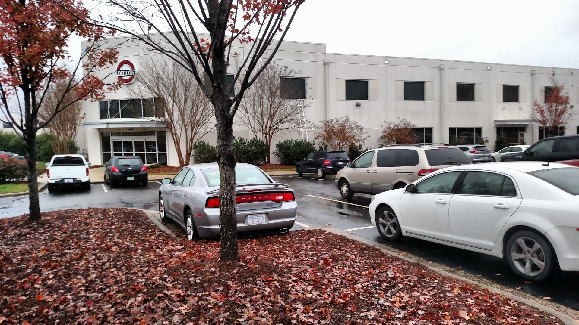 Parking lot outside a white office building with several cars and red autumn leaves on the ground.