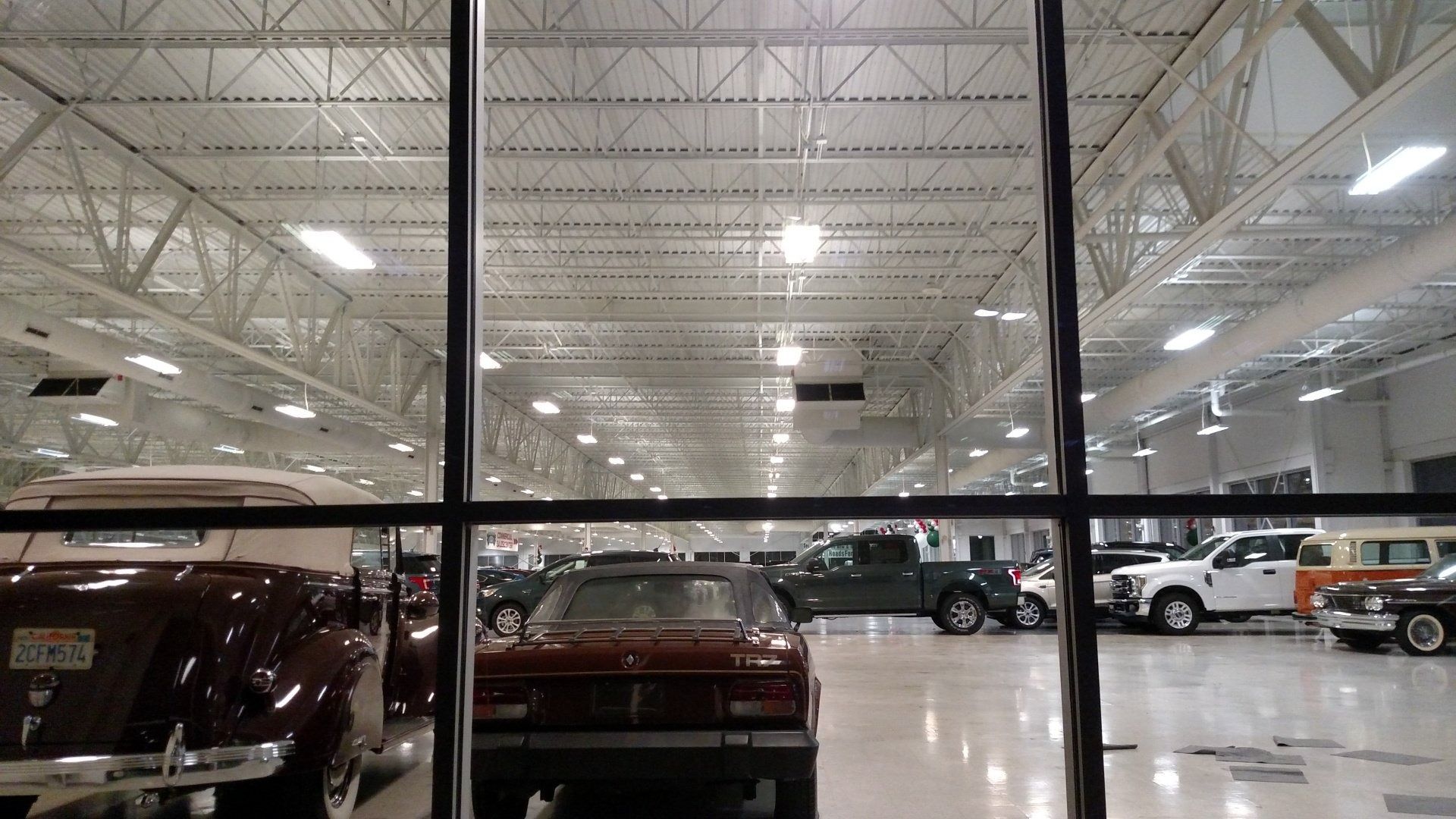 Indoor car showroom with several parked cars under bright lights, viewed through a window.