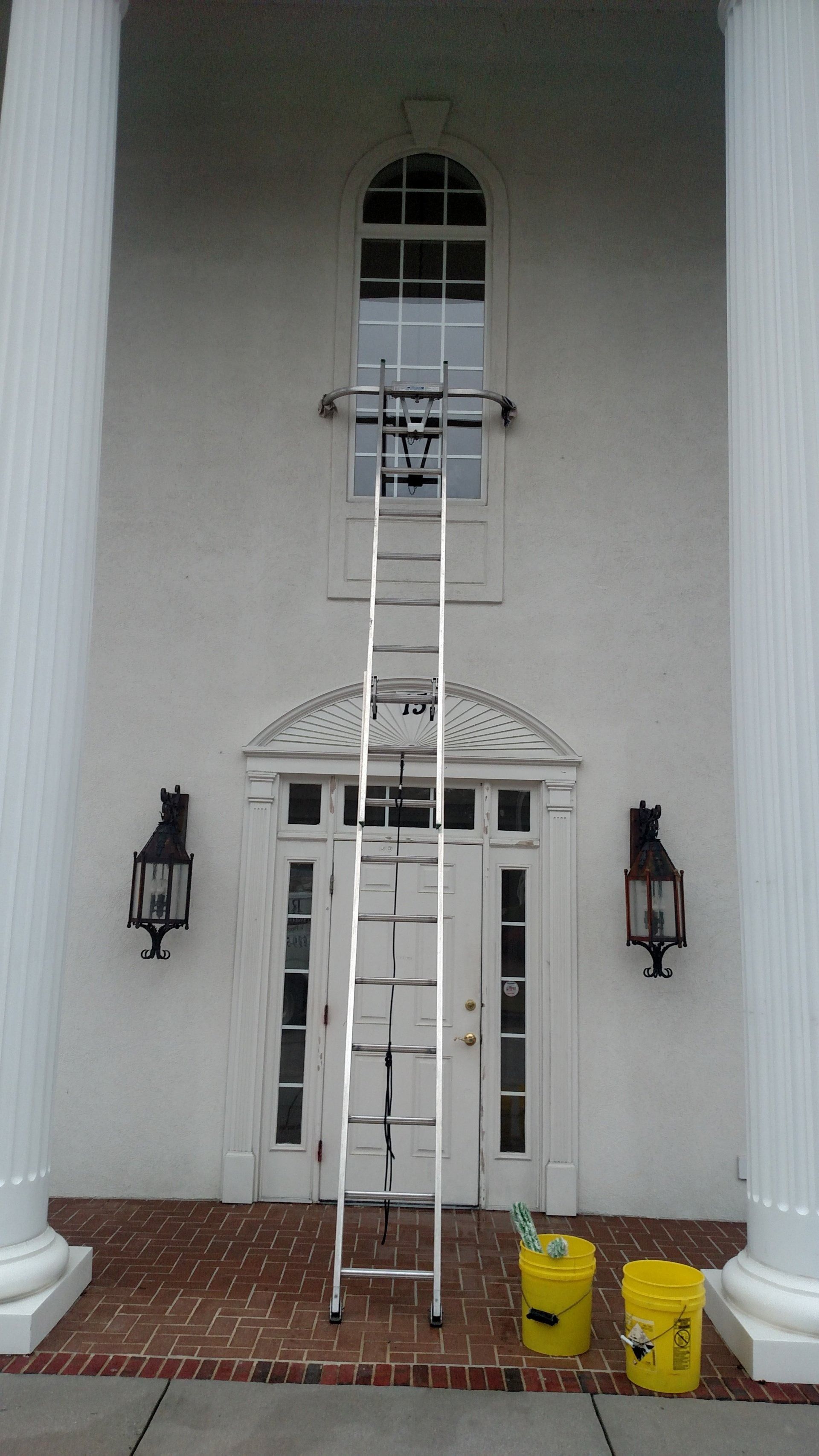 A long ladder leans against a white house facade above a front door with lanterns and yellow buckets below.