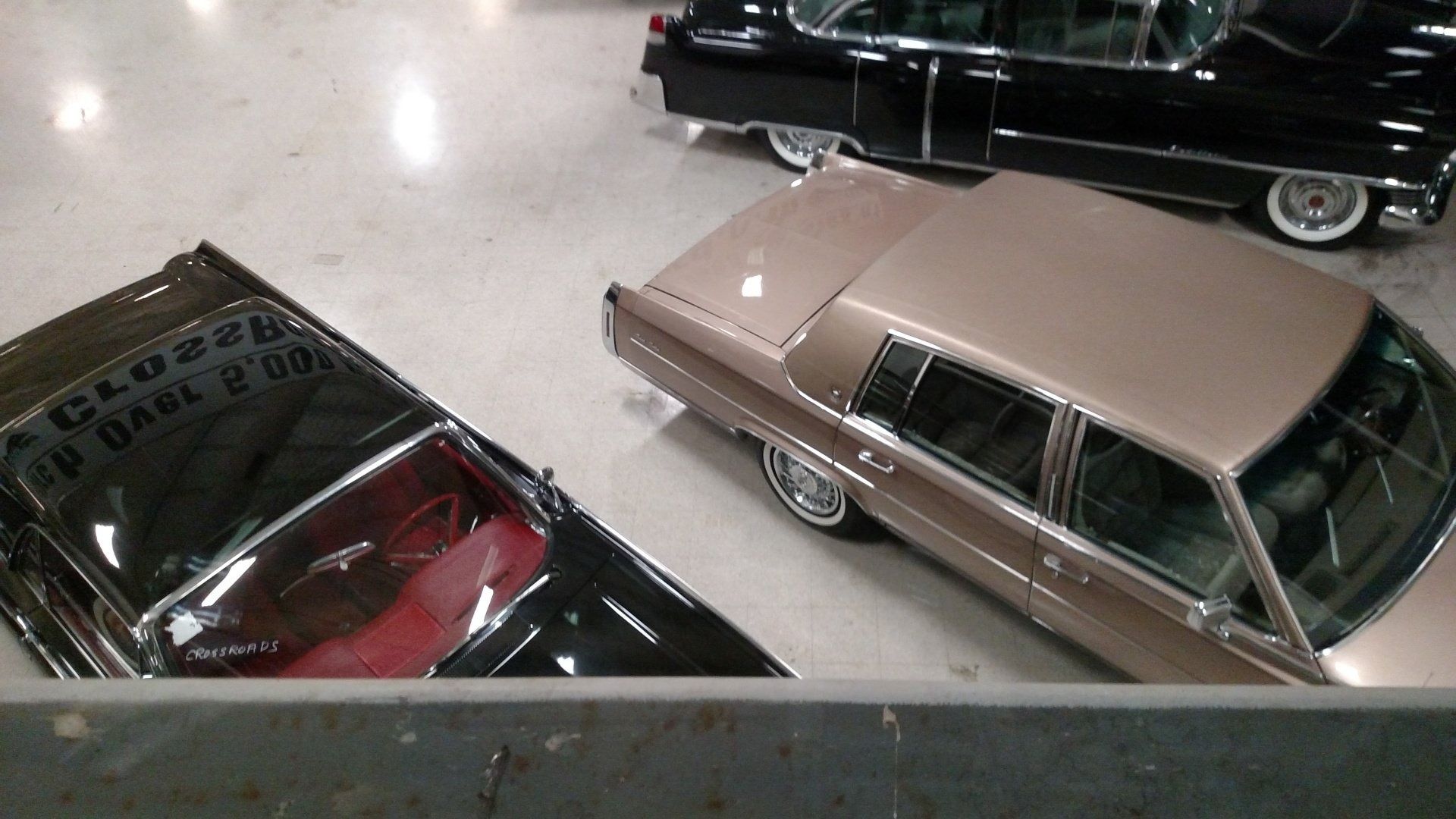 Three classic cars displayed indoors on a polished showroom floor, including a tan sedan and black cars.