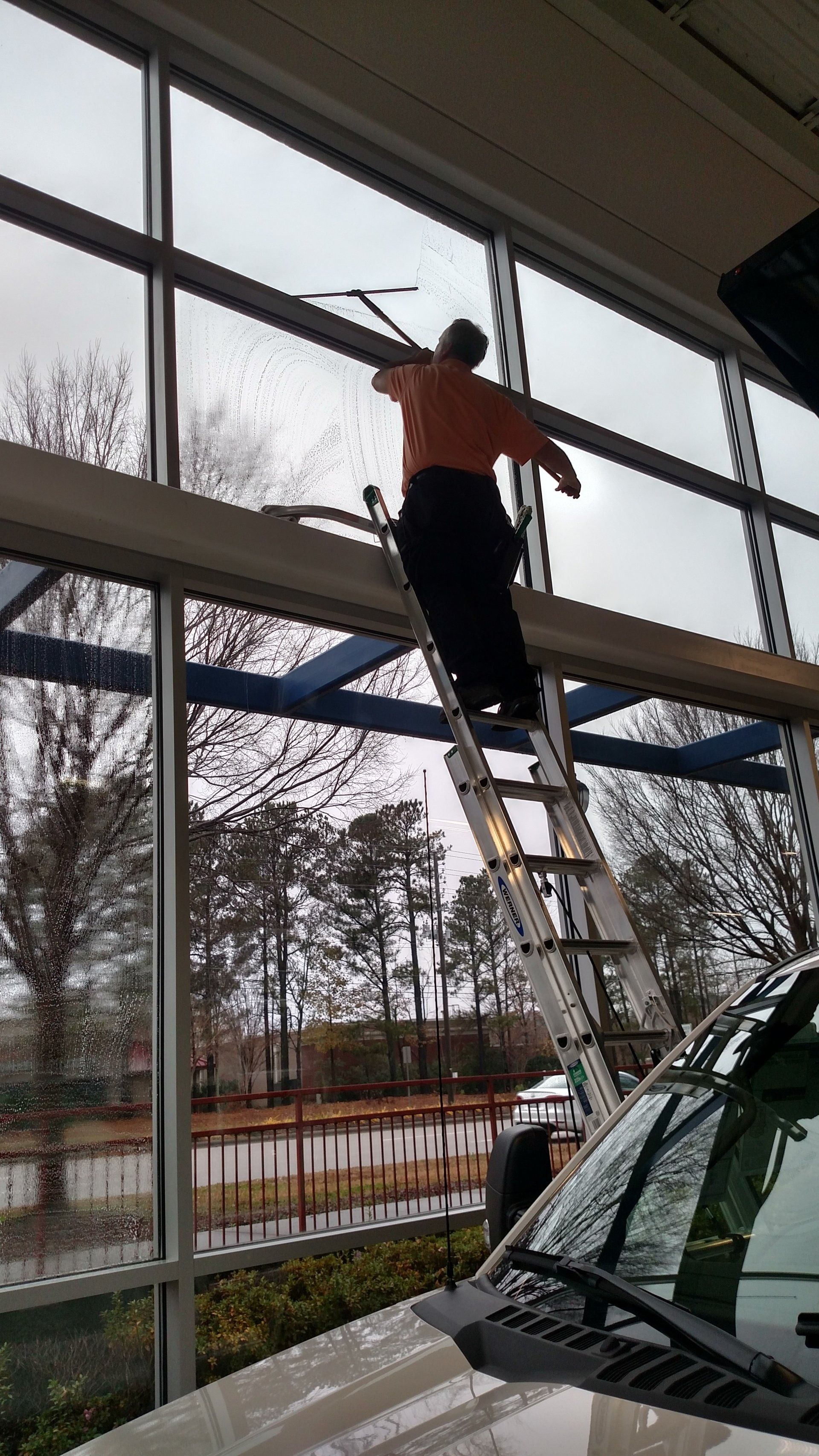Person on a ladder working near large windows inside a bright garage, with a car below.
