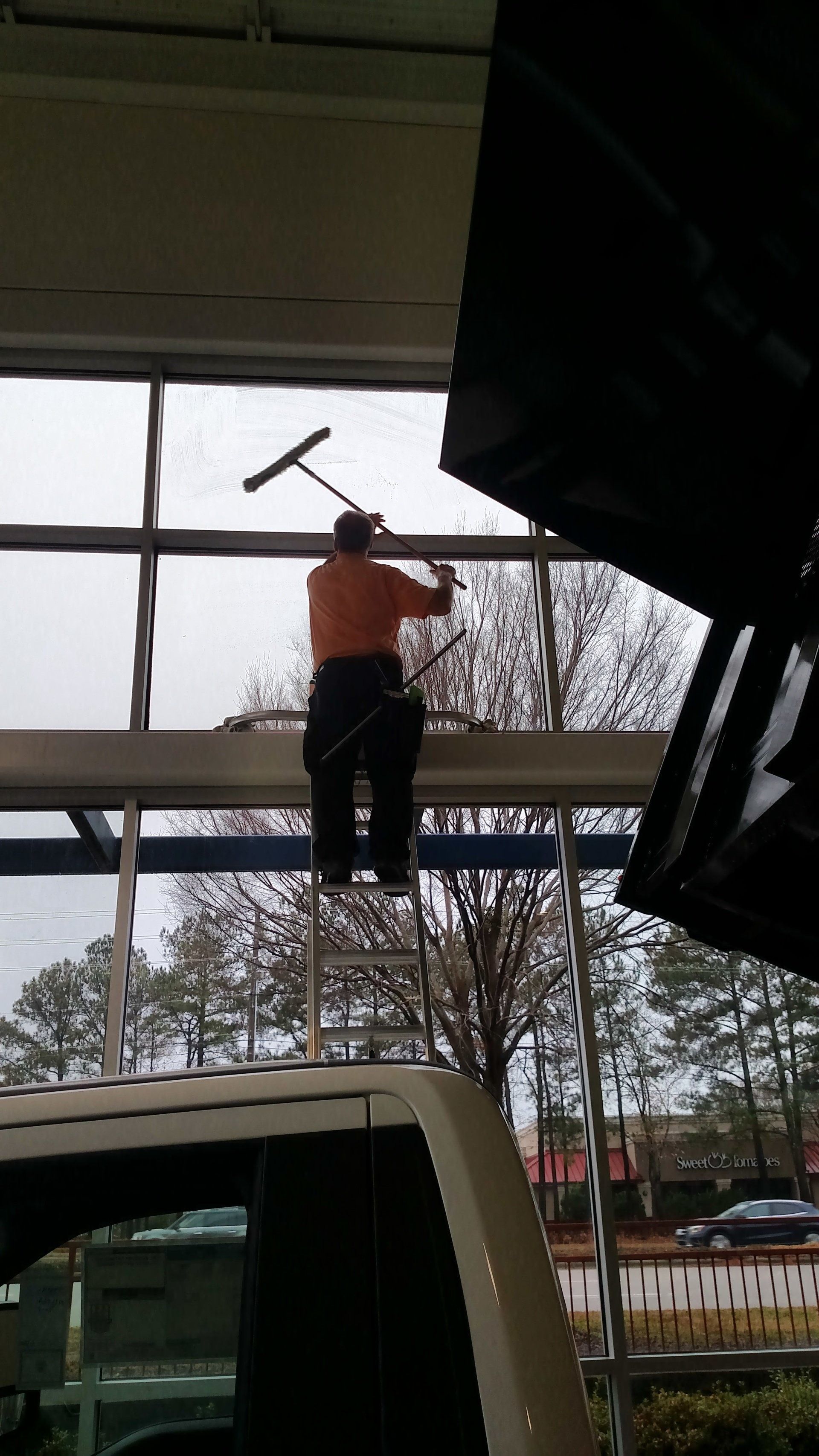 Person in an orange shirt power-washing a window on a raised platform beside a parked van.