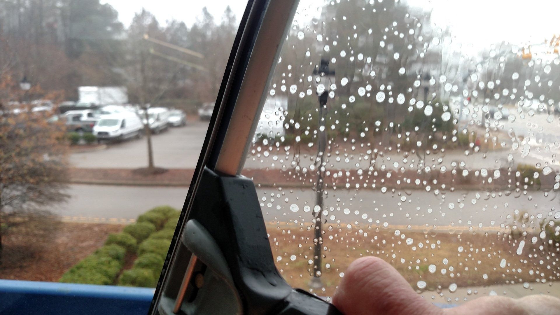 Rainy parking lot seen through a car window with raindrops on the glass and a hand near the frame