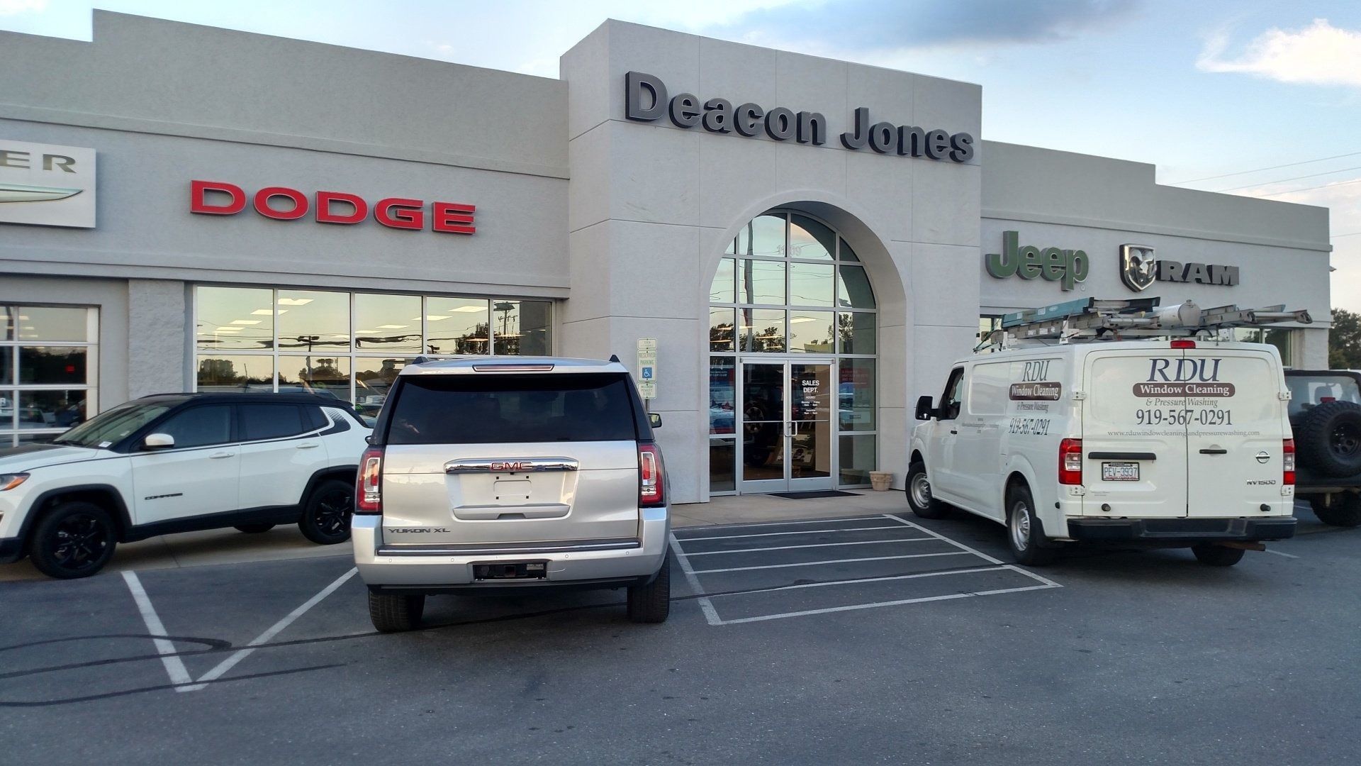 Car dealership storefront with Dodge signage, several parked vehicles, and a van in front of the entrance