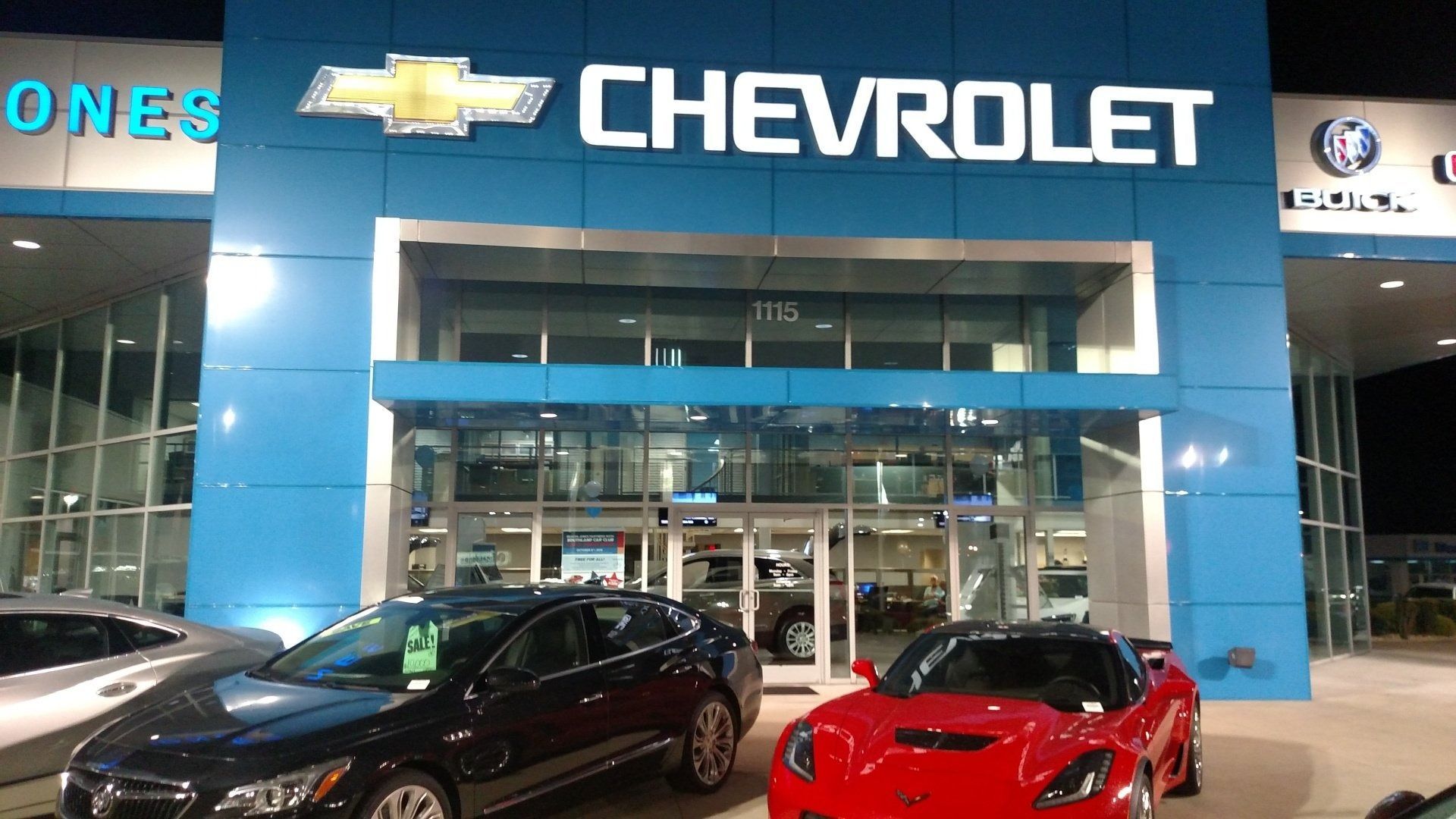 Chevrolet dealership storefront at night with two cars parked outside, including a red sports car