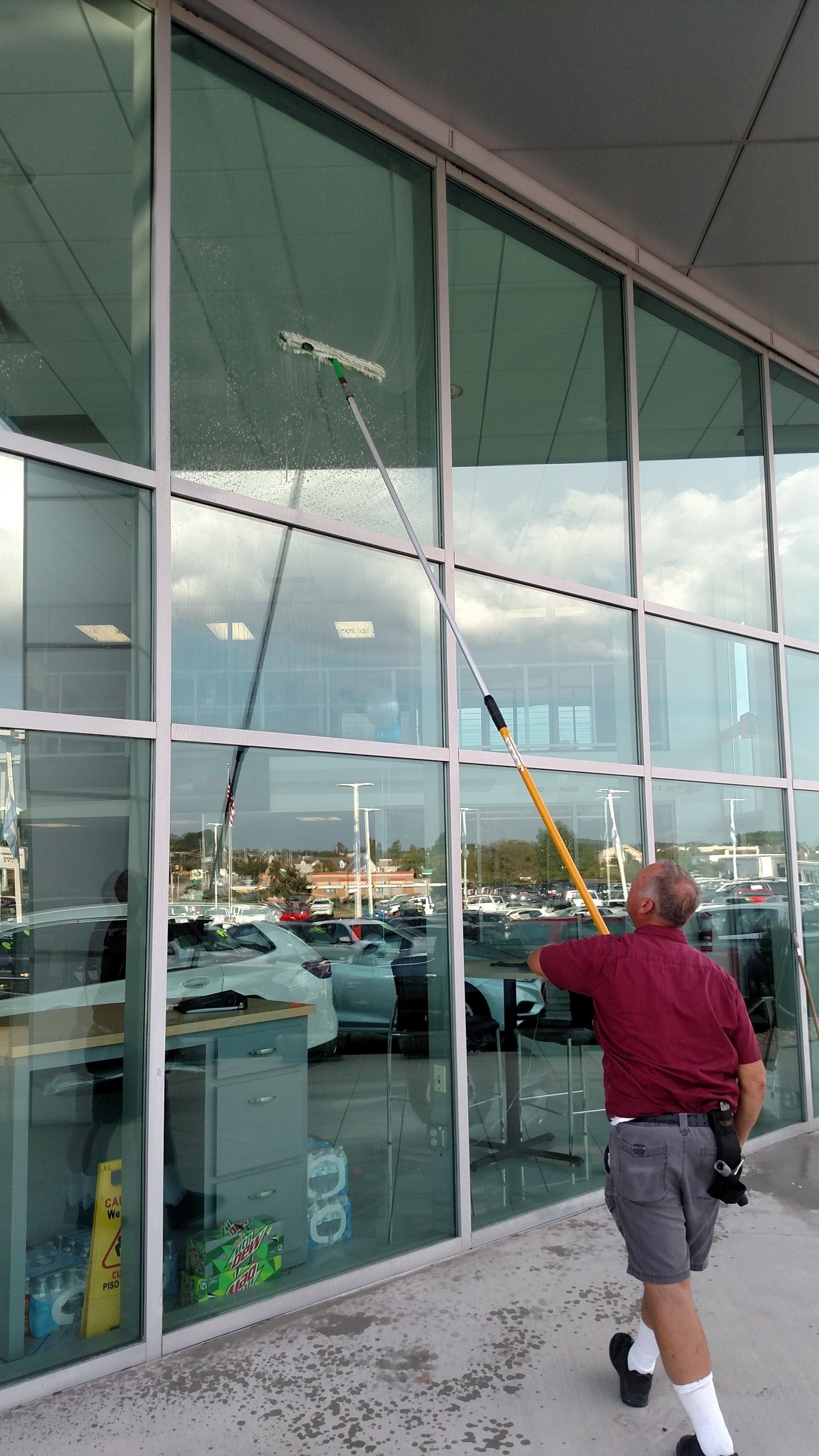 Person using a long-handled squeegee to clean tall glass windows outside a building.
