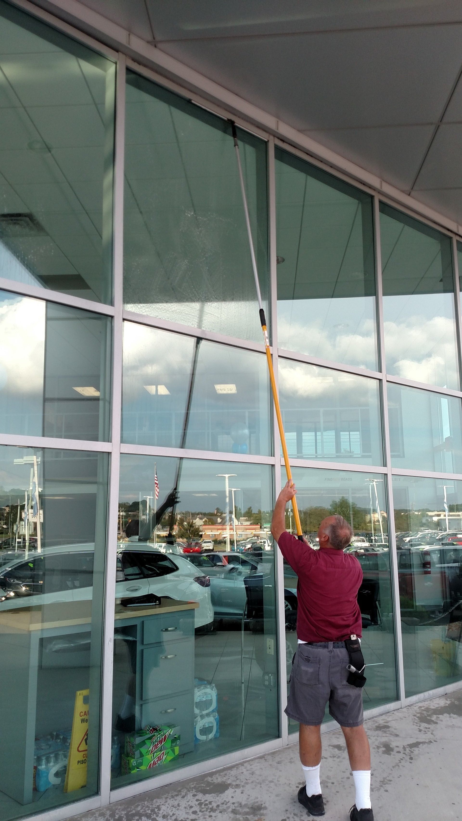 Person in a red shirt reaching up to clean large glass windows outside a modern building.