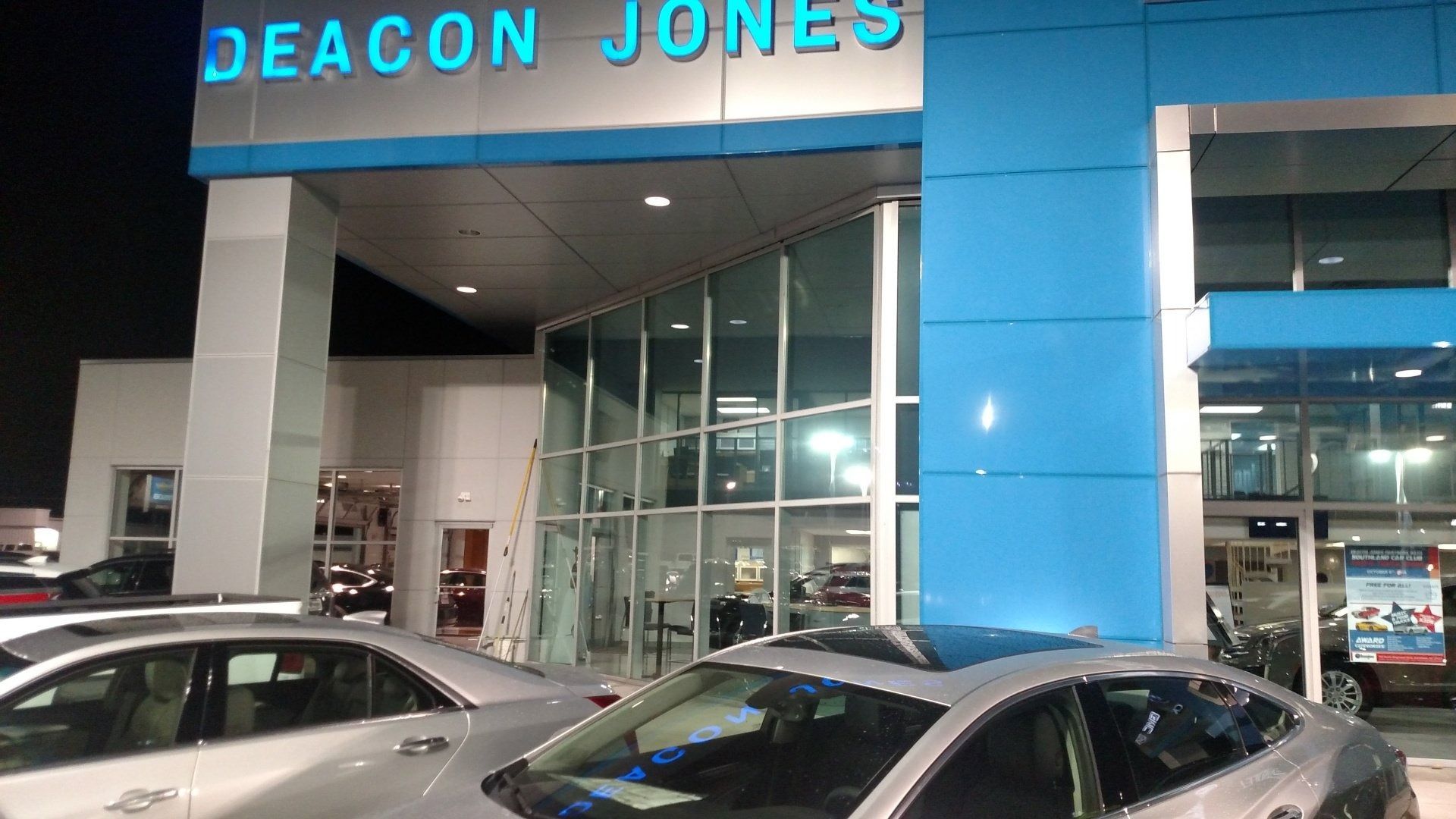 Nighttime exterior of a Deacon Jones car dealership with parked cars and a glass entrance.