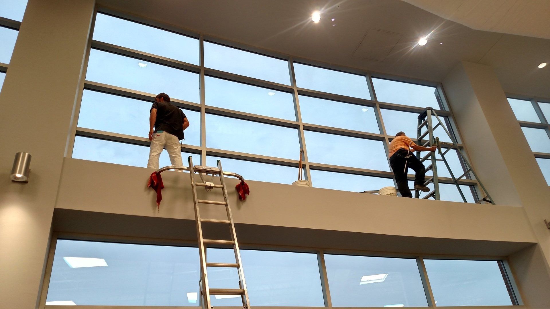 Two workers on a ladder and mezzanine inside a modern building, installing or repairing windows.