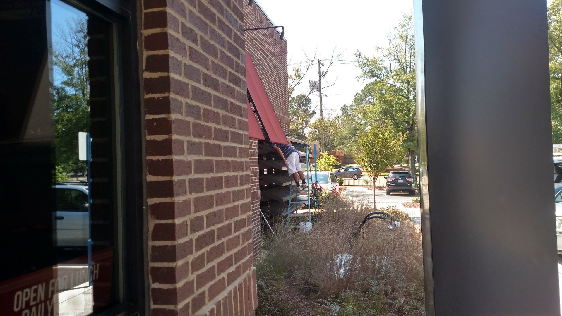 Brick storefront corner with red awning and sidewalk plants, looking out to a street and parked cars