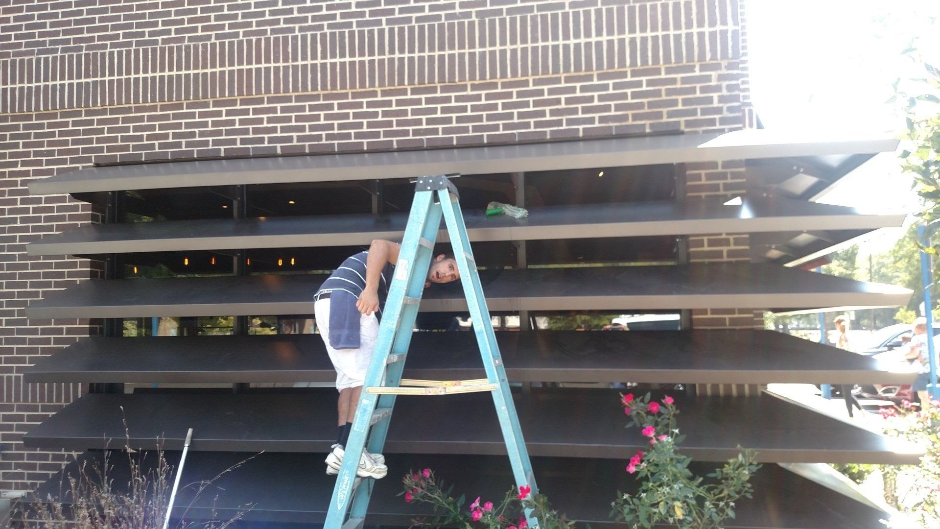 Person on a blue ladder beside a brick house, working on horizontal wooden slats with flowers below