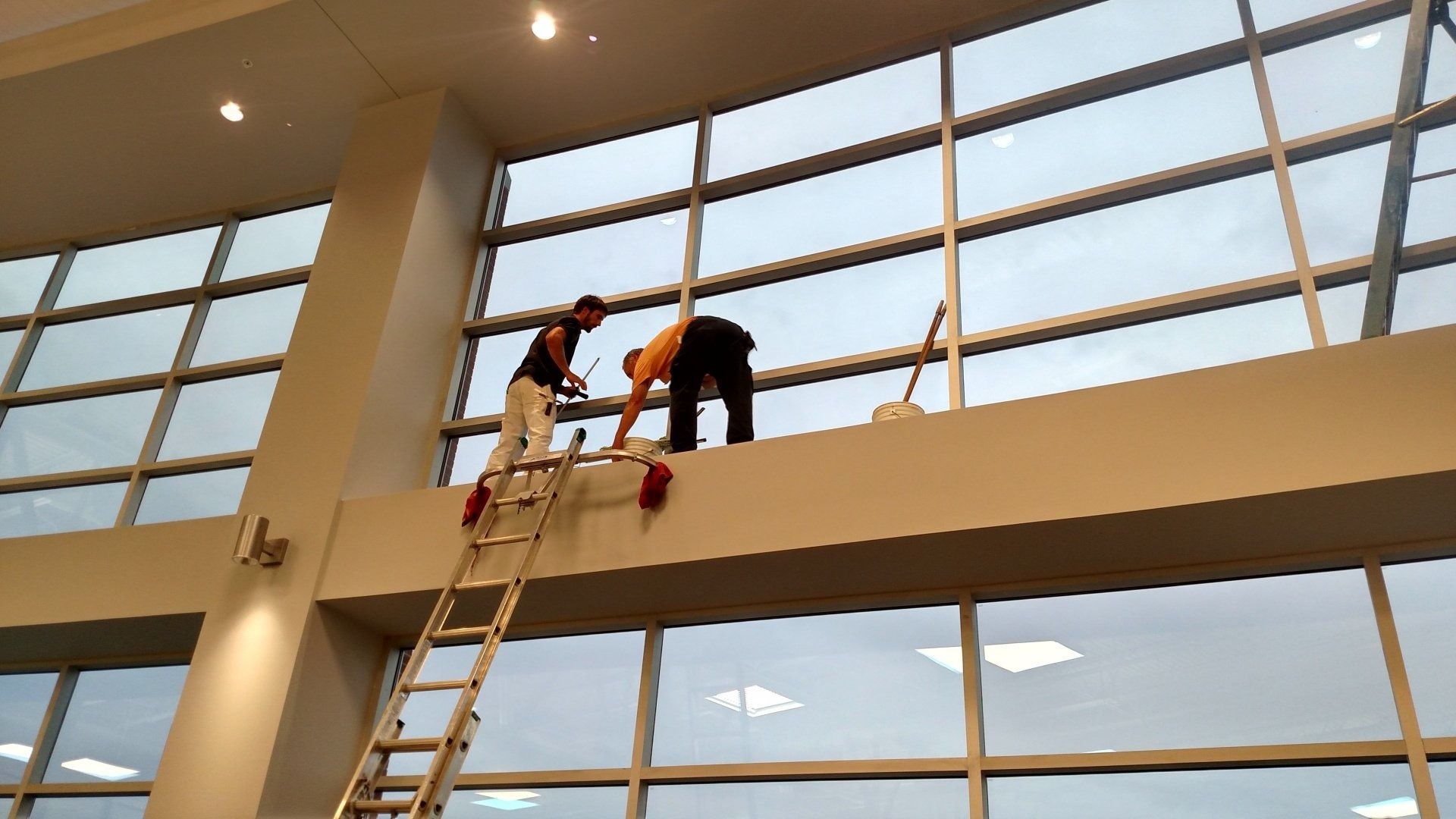 Two workers on a ladder repairing windows in a large atrium with tall glass walls