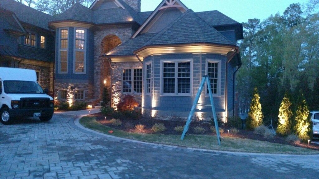 Suburban brick house at dusk with lit windows, driveway, and a ladder leaning near the front entrance.